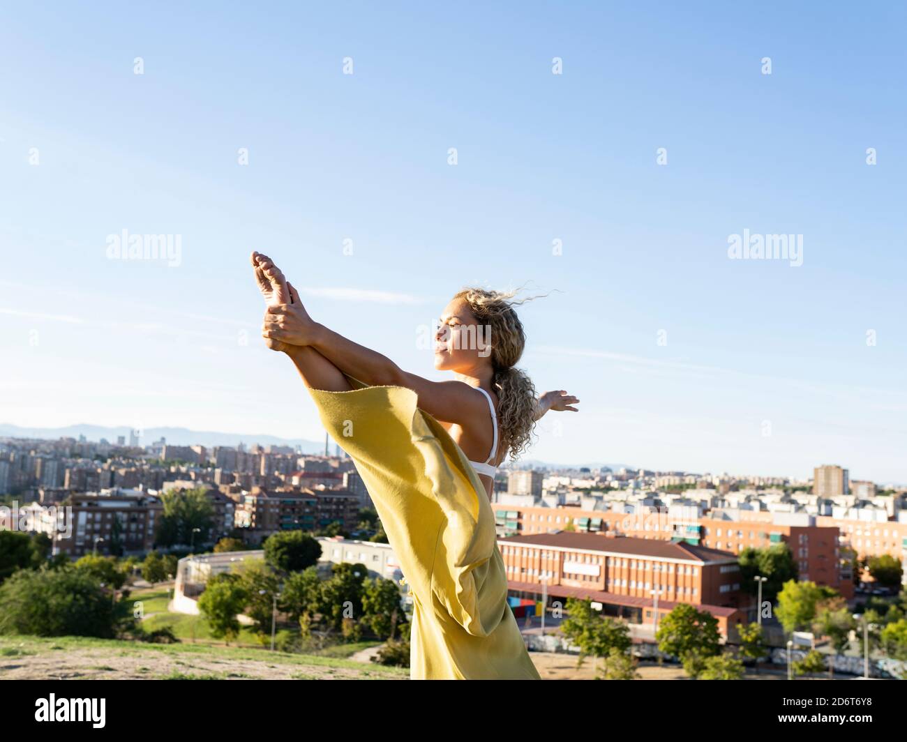 Schlanke, flexible Frau in lockerer Hose mit verlängerter Hand Big Toe Pose beim Üben Yoga auf sonnigen Hügel gegen Städtische Umwelt Stockfoto