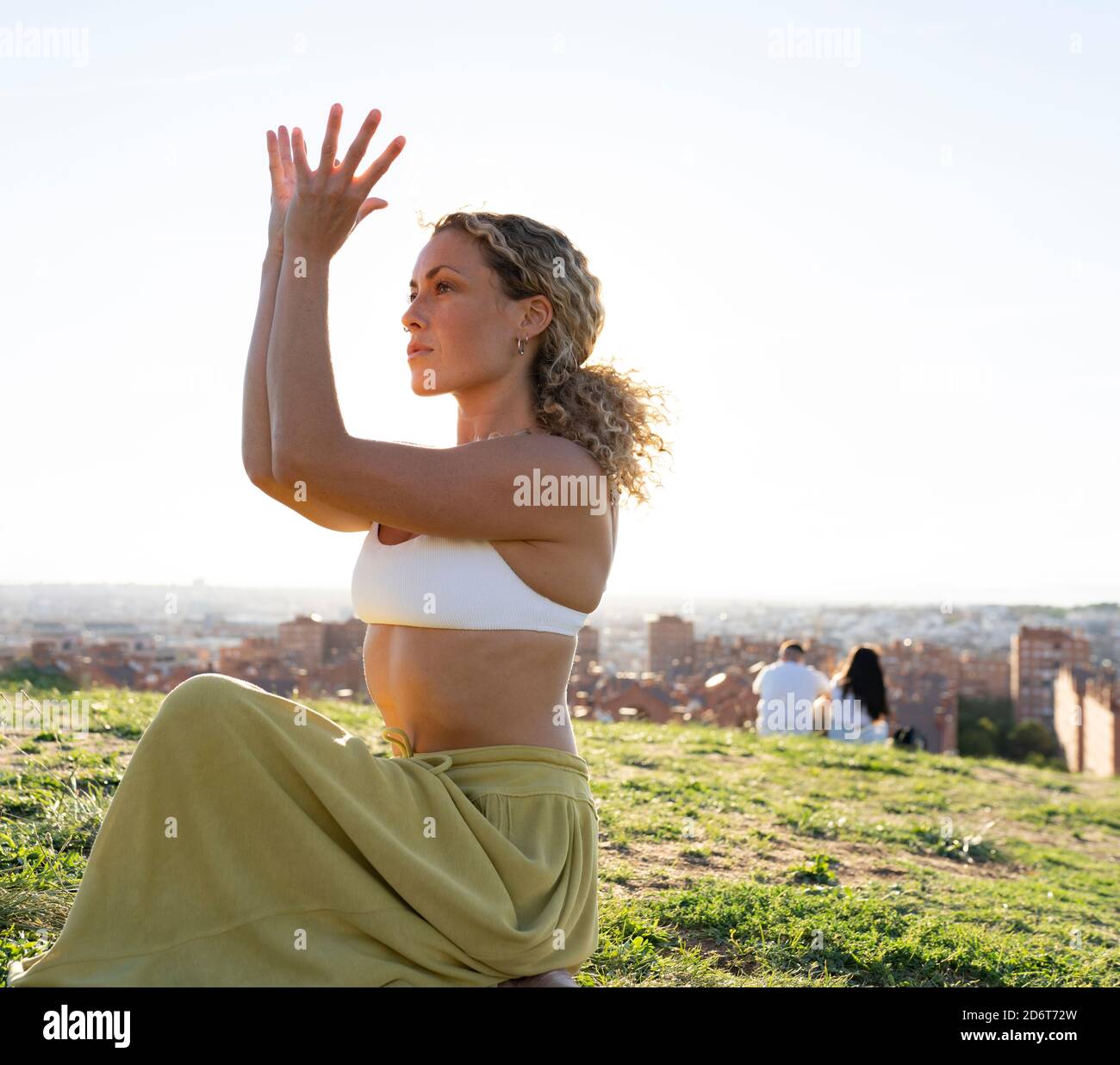 Ganzkörper barfuß weiblich sitzen in Kuh Gesicht Pose und Das Gesicht hinter den Armen verstecken, während Sie Yoga auf einem sonnigen Hügel praktizieren Gegen die Stadtlandschaft Stockfoto