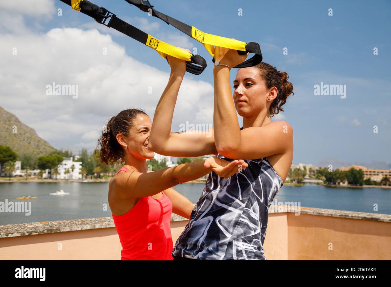 Expressive weibliche Trainerin unterstützt die konzentrierte Sportlerin beim Training auf der Terrasse im Sommer bei Übungen mit TRX-Trägern Stockfoto