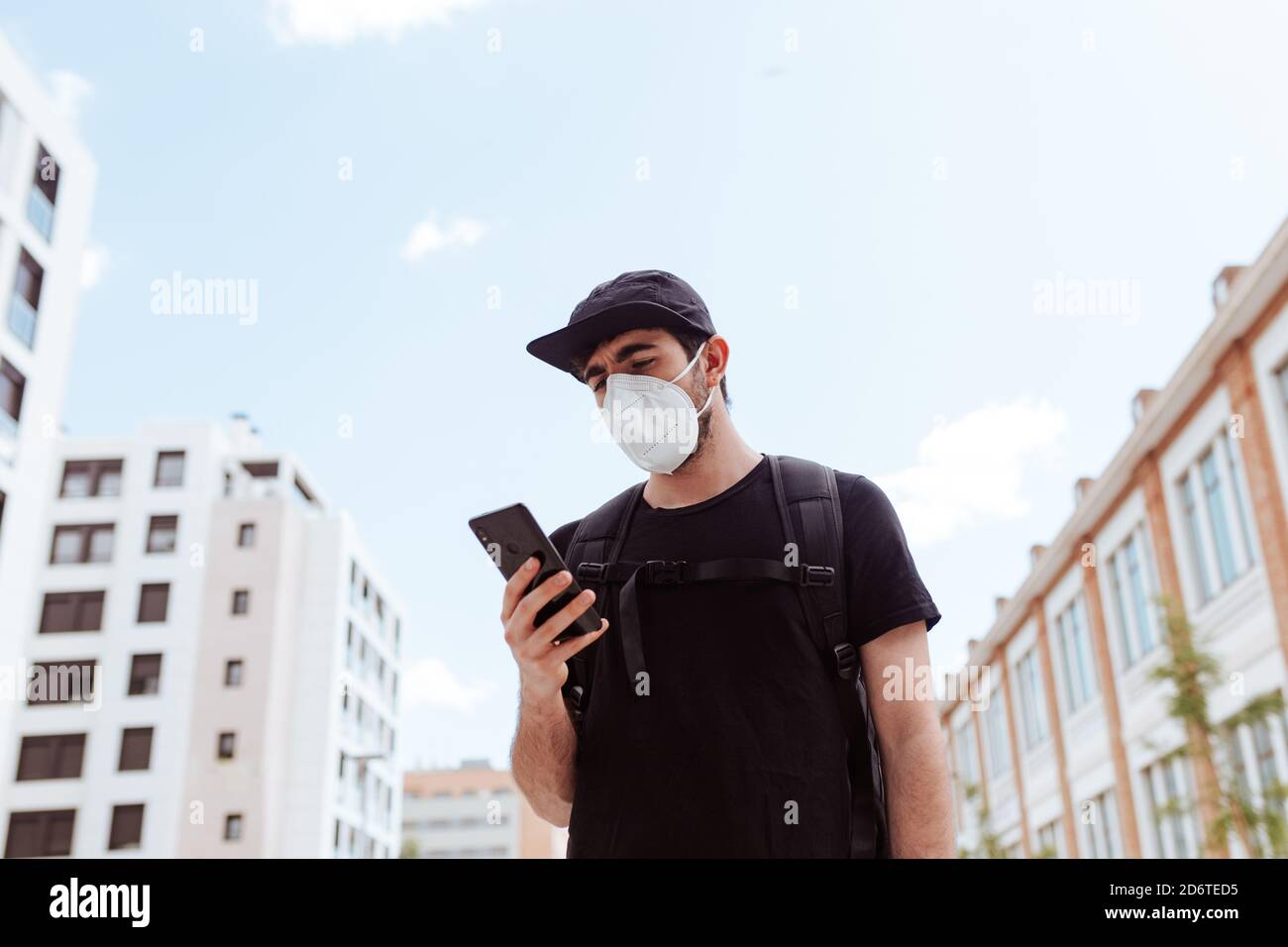 Von unten von unerkennbarem Männchen in Gesichtsmaske und Mütze Surfen im Internet auf Handy unter Himmel während COVID 19 Pandemie Stockfoto