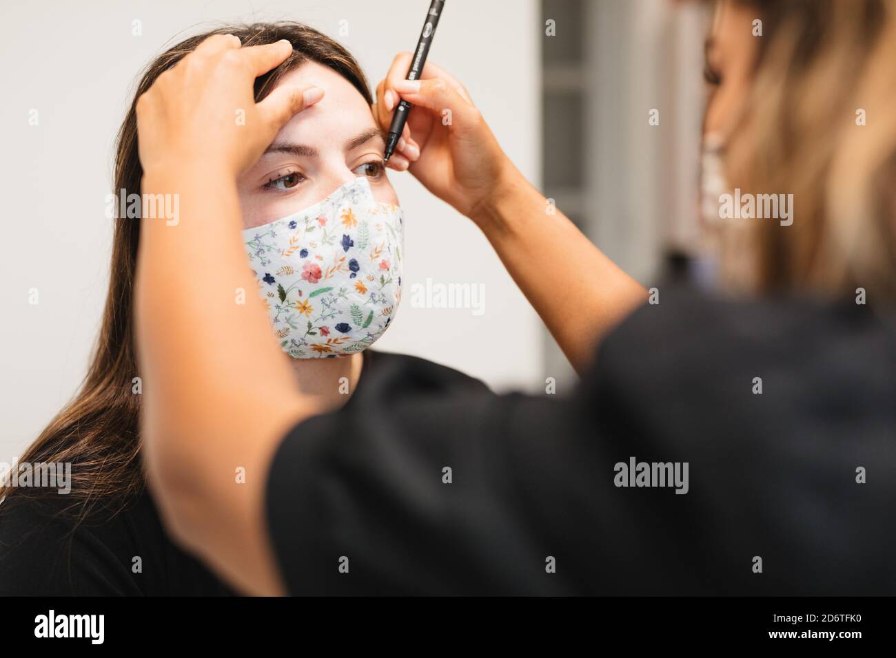 Nicht erkennbare weibliche Visagiste Anwendung Eyeliner auf Gesicht der jungen Frau in medizinische Maske vor Wimpern Verlängerung Stockfoto