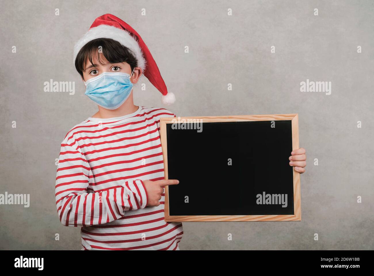 Frohe Weihnachten, lustiges Kind mit medizinischer Maske, die eine Tafel hält Grauer Hintergrund Stockfoto