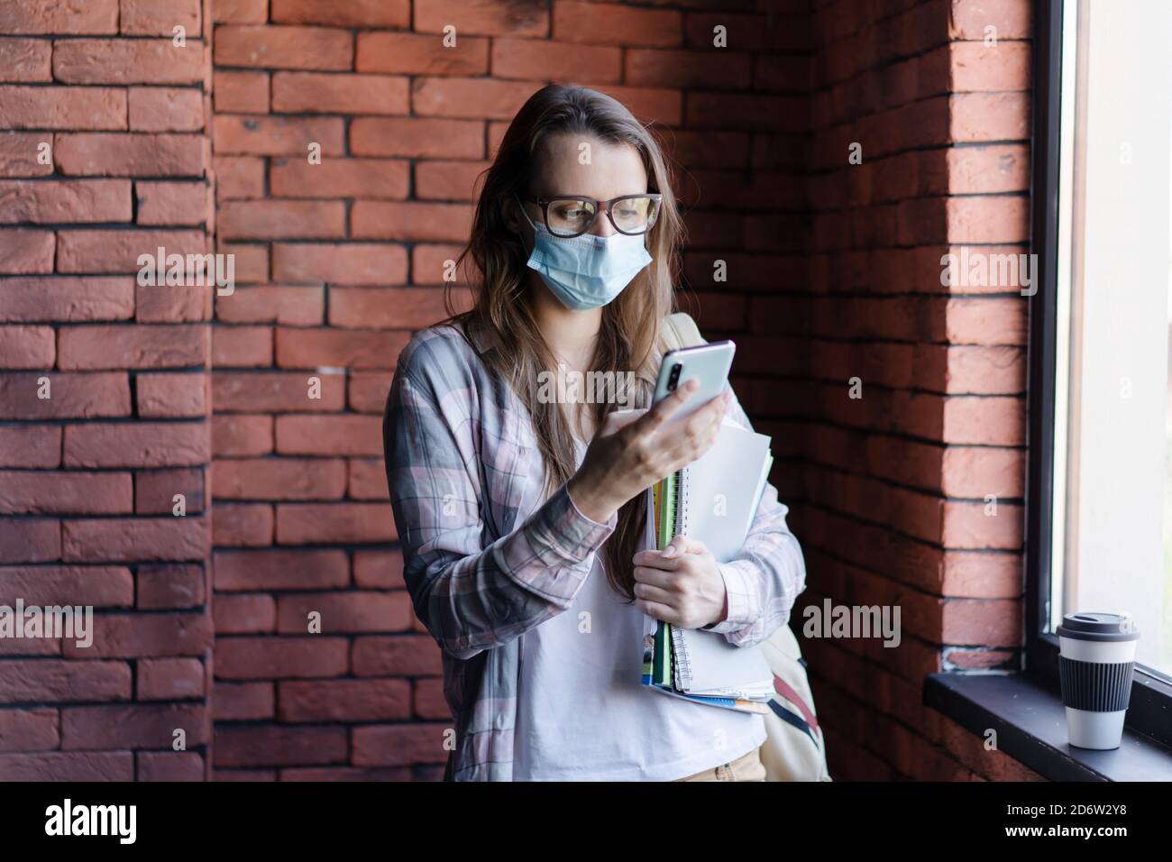 Studentin in einer schützenden medizinischen Maske auf ihrem Gesicht schaut auf das Telefon. Stockfoto
