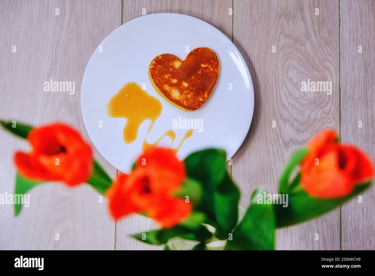 Herzförmiger Pfannkuchen und rote Tulpen. Backen Favoriten und Blumen am Valentinstag. Ein Teller mit Pfannkuchen, Honig und Sirup auf einem Holztisch. Ein Geschenk für Stockfoto