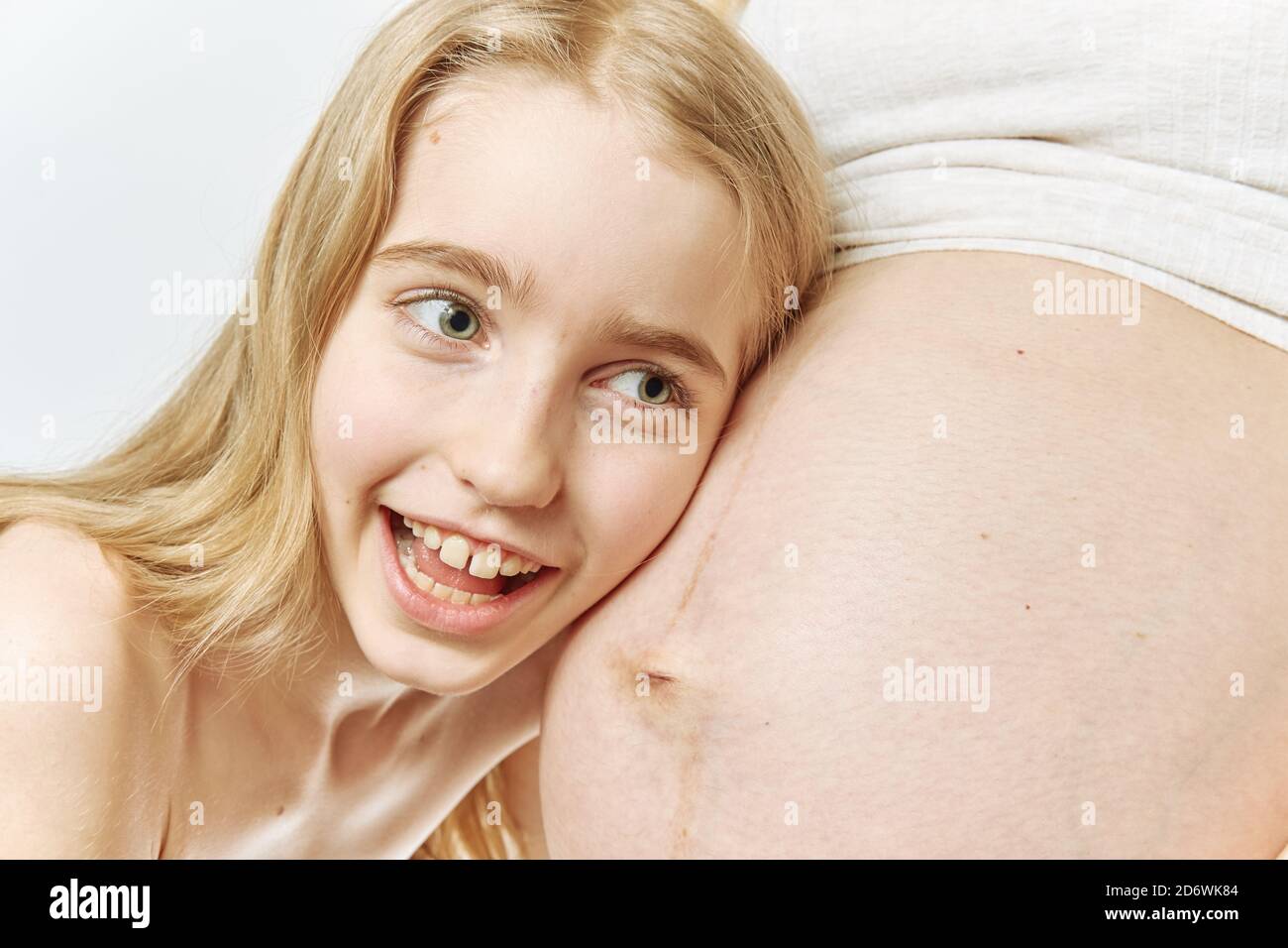 Ein blondes kaukasisches Mädchen umarmt sich und lauscht dem Bauch ihrer schwangeren Mutter. Stockfoto