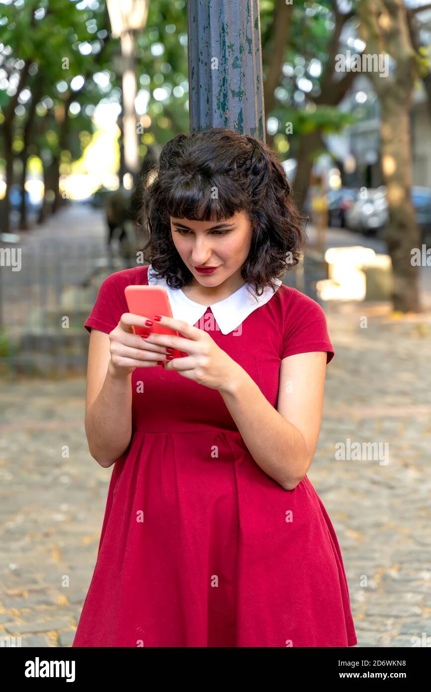 Eine schöne Frau in einem roten Kleid mit Ihrem Smartphone, während neben einem Lamp Post steht auf einem sonnigen Tag. Stockfoto