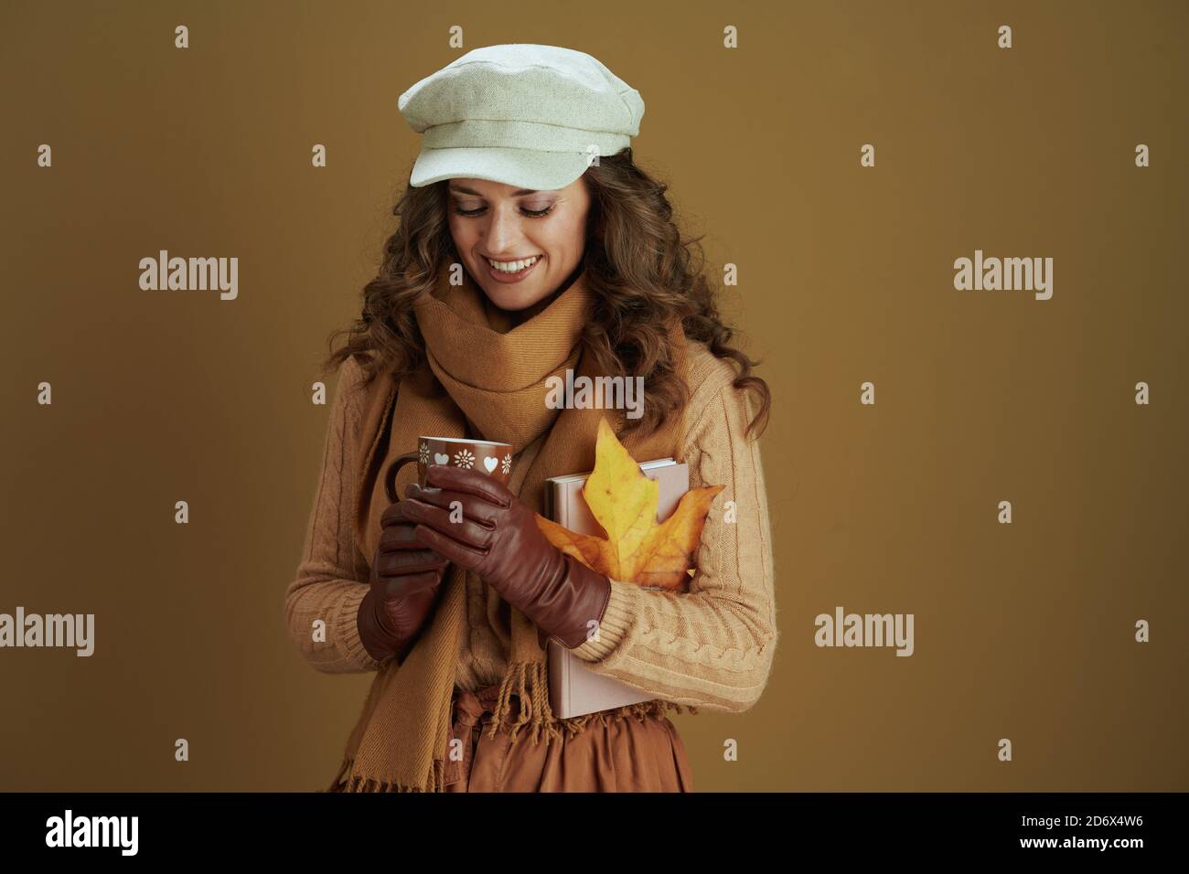 Hallo Herbst. Happy trendy 40 Jahre alte Hausfrau im Schal mit gelben Herbst Ahornblatt, Buch, Lederhandschuhe und Becher auf braunem Hintergrund. Stockfoto