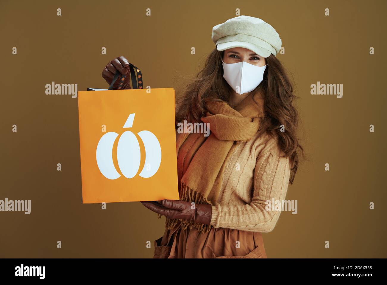Leben während covid-19 Pandemie. Lächelnd elegante Frau in Schal mit weißen medizinischen Maske und Papier Einkaufstasche auf braunem Hintergrund. Stockfoto