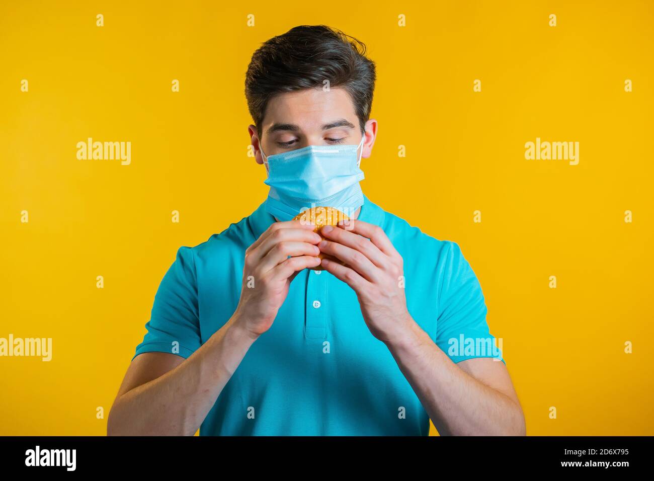 Junger Mann in schützende medizinische Maske essen Burger auf gelben Studio-Hintergrund. Guy macht Spaß, zeigt, wie man während einer Epidemie lebt. Stockfoto