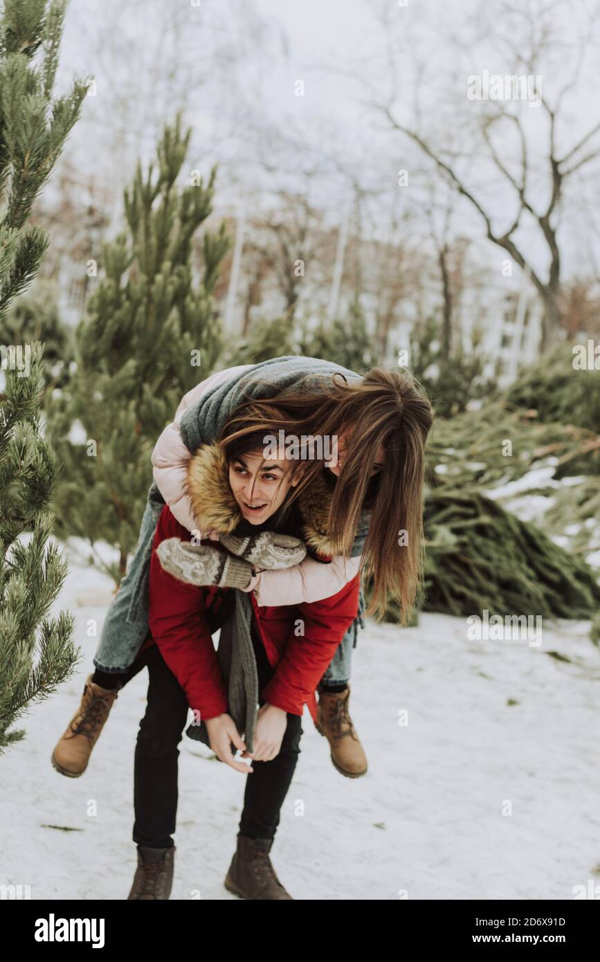 Ein junges Paar macht sich im Winter vor dem Hintergrund des Christbaummarktes in der Stadt lustig. Eine junge Frau auf dem Rücken ihres Freundes lacht und lächelt. Stockfoto