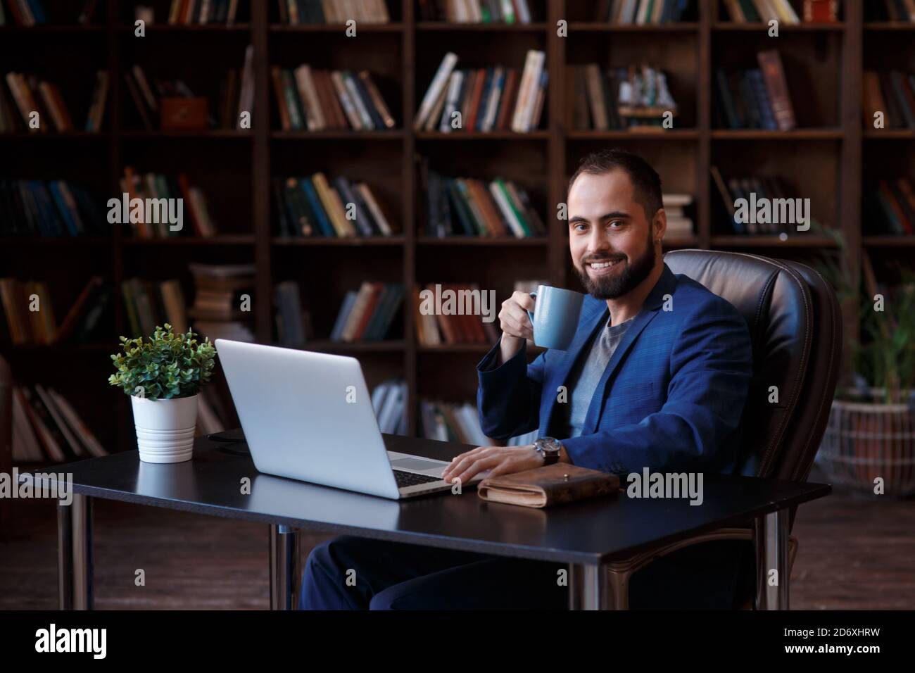 Genießen Sie frischen Kaffee bei der Arbeit. Selbstbewusster junger Mann in eleganter Freizeitkleidung, der am Laptop arbeitet und ein heißes Getränk trinkt, während er bei seiner Arbeit sitzt Stockfoto