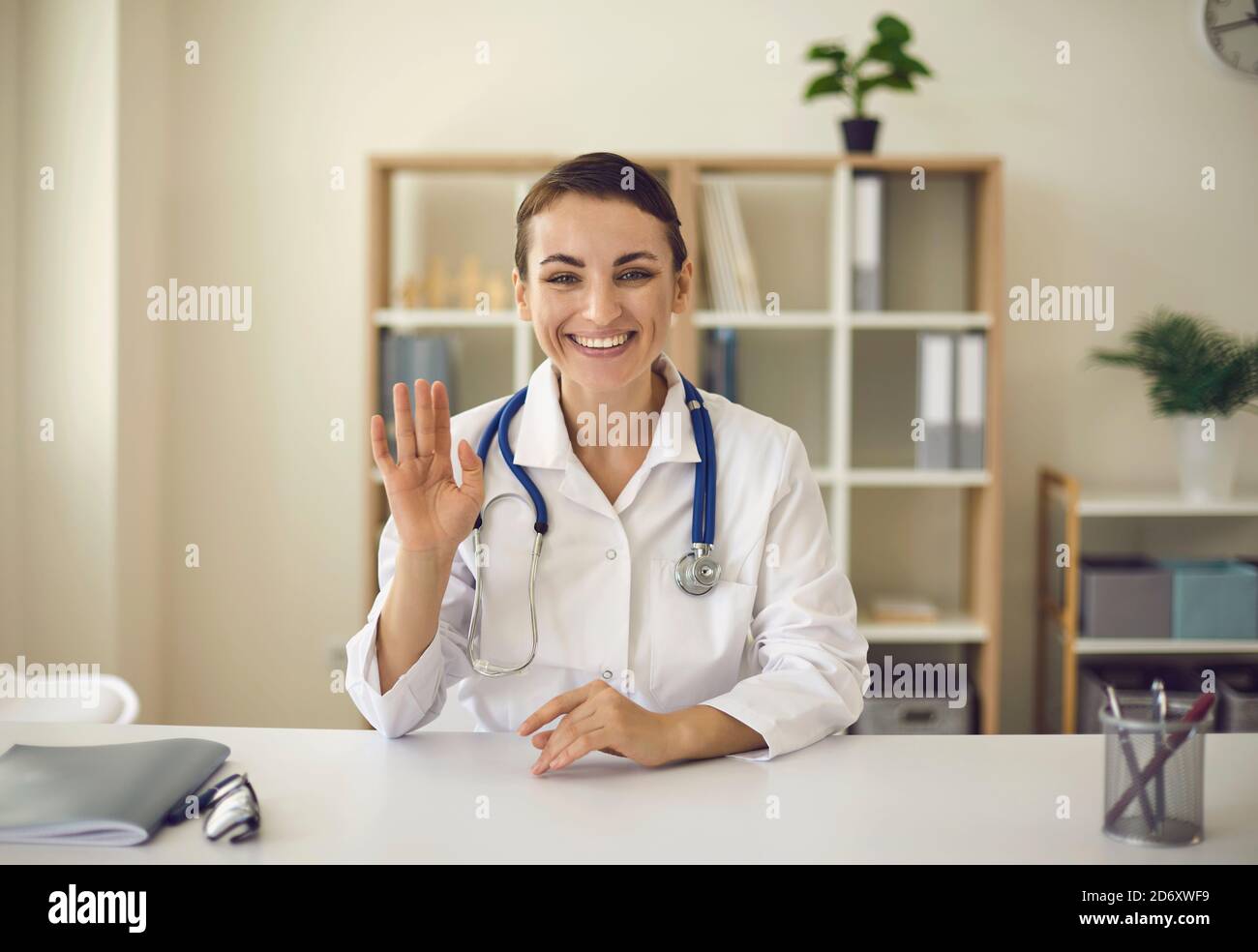 Junge lächelnde Ärztin, die Kamera anschaut und jemanden grüßt Online Stockfoto