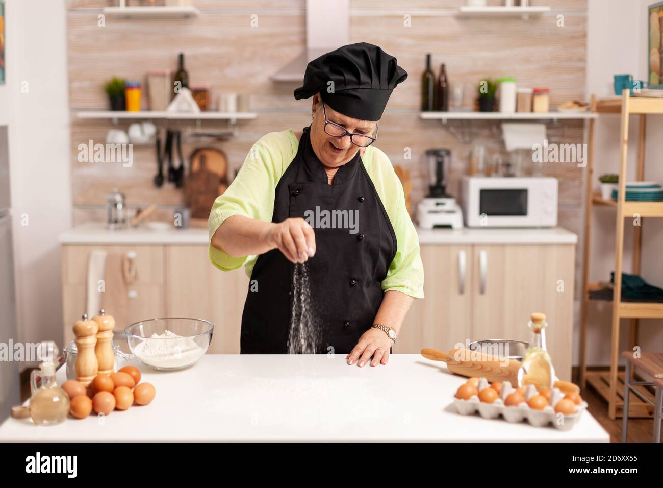 Die Chefköchin lächelte, während sie Pizza streute und Mehl auf den Küchentisch gab. Glücklicher älterer Koch mit einheitlicher Streuselung, sieben rohe Zutaten von Hand. Stockfoto