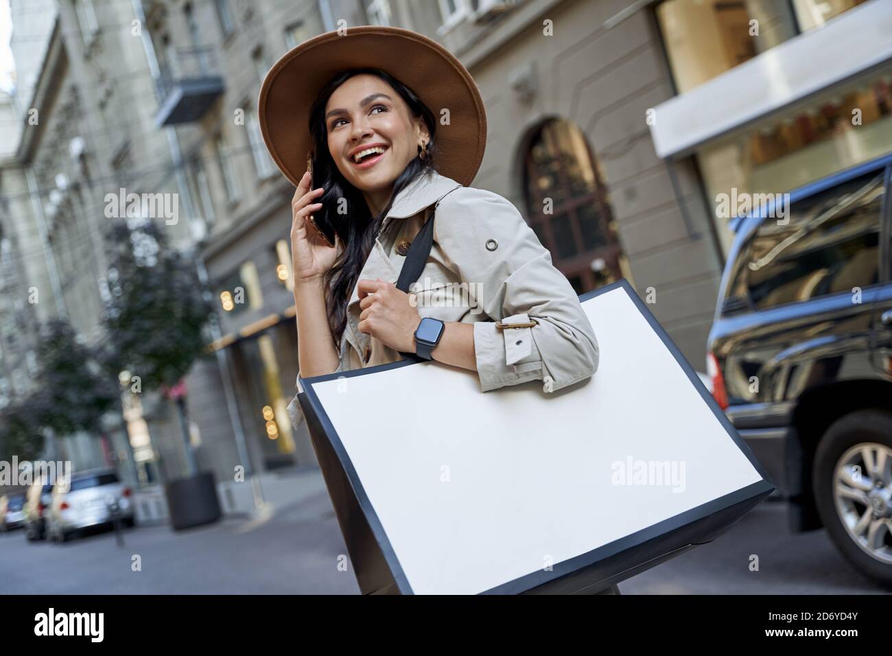 Einkaufen macht uns glücklich. Junge schöne aufgeregt Frau in Hut mit Einkaufstasche reden auf dem Smartphone, Blick zur Seite und lächeln, während Sie auf den Straßen der Stadt. Mode, Menschen Lifestyle-Konzept Stockfoto