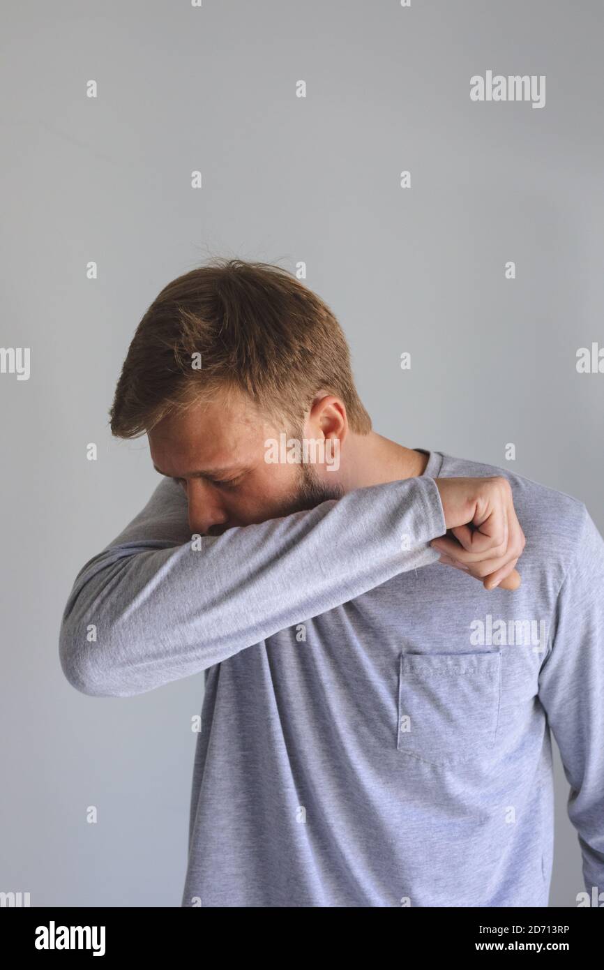 Kaukasischer Mann verbringt Zeit zu Hause, krank, hustet niesen, bedeckt sein Gesicht mit Ellbogen auf grauem Hintergrund. Soziale Distanzierung während Covid 19 Stockfoto