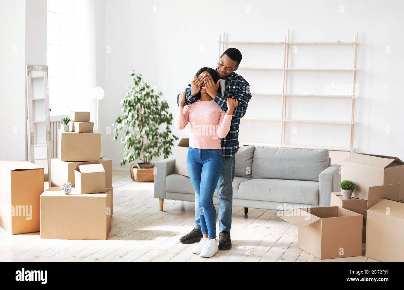 Umzugskonzept. Millennial schwarzer Mann schließt die Augen seiner Freundin in ihrem Haus mit Kartons Stockfoto