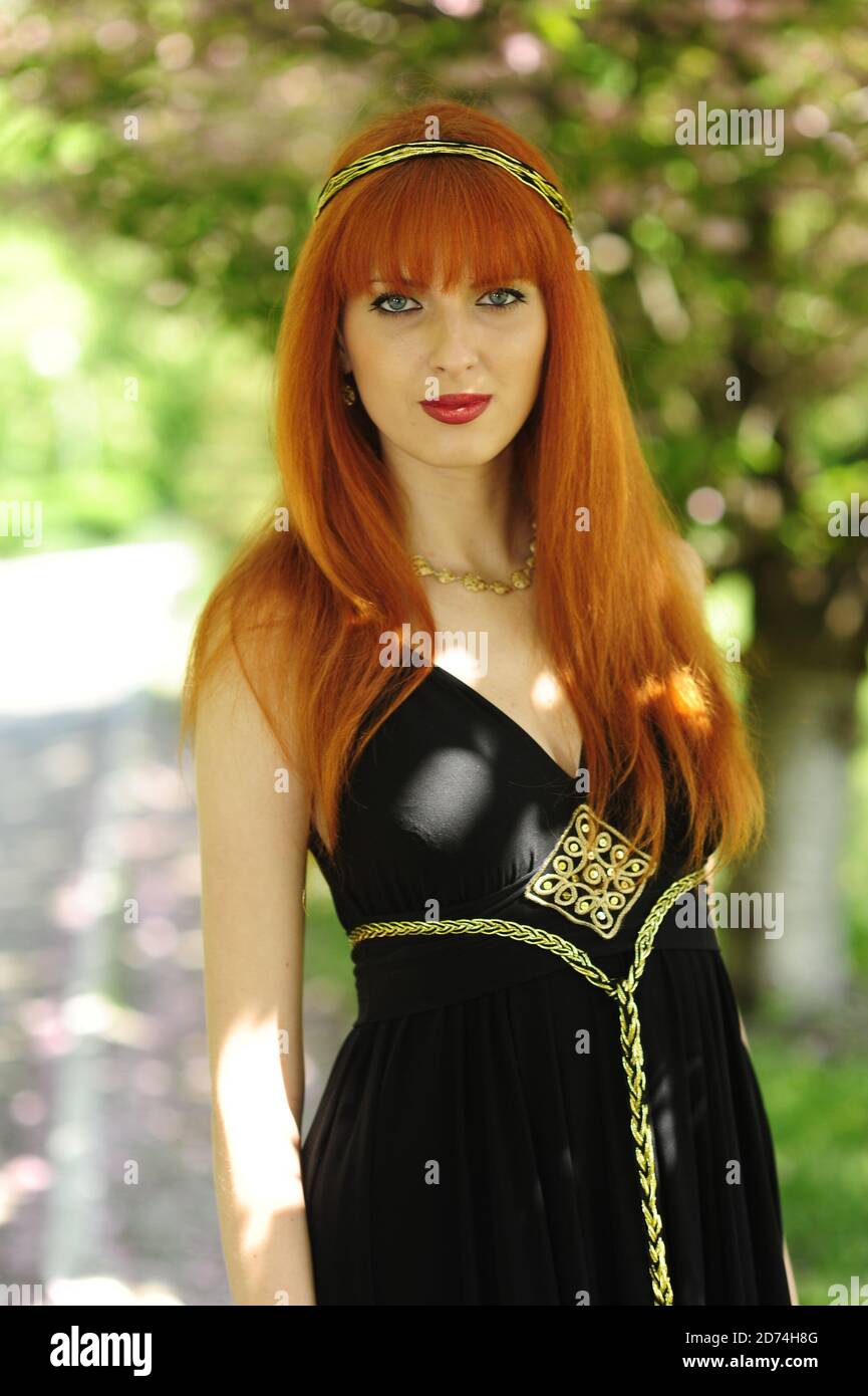 Junge schöne Frau mit roten Haaren in einem schwarzen langen Kleiden Sie sich in einem Frühlingsgarten auf einem Hintergrund der Blüte sakura Stockfoto