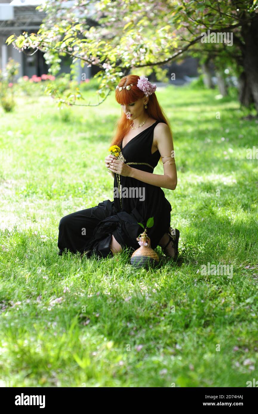 Junge schöne Frau mit roten Haaren in einem schwarzen langen Kleiden Sie sich in einem Frühlingsgarten auf einem Hintergrund der Blüte sakura Stockfoto