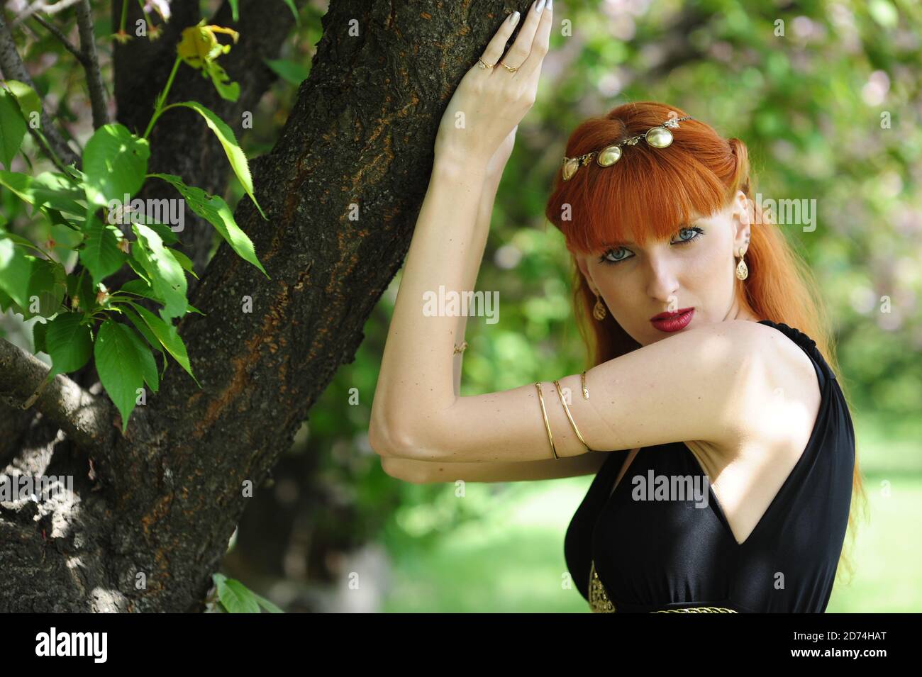 Junge schöne Frau mit roten Haaren in einem schwarzen langen Kleiden Sie sich in einem Frühlingsgarten auf einem Hintergrund der Blüte sakura Stockfoto