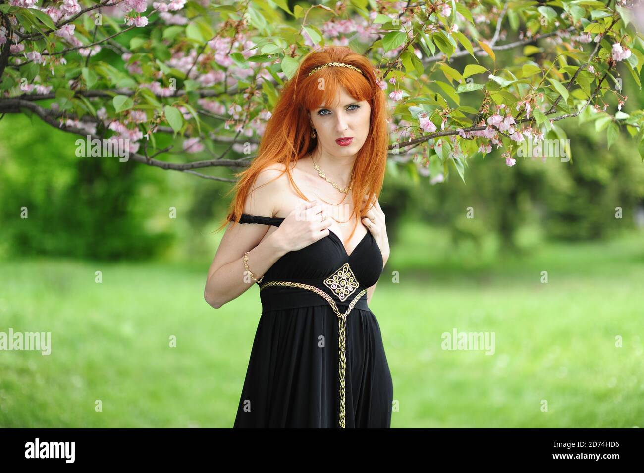 Junge schöne Frau mit roten Haaren in einem schwarzen langen Kleiden Sie sich in einem Frühlingsgarten auf einem Hintergrund der Blüte sakura Stockfoto