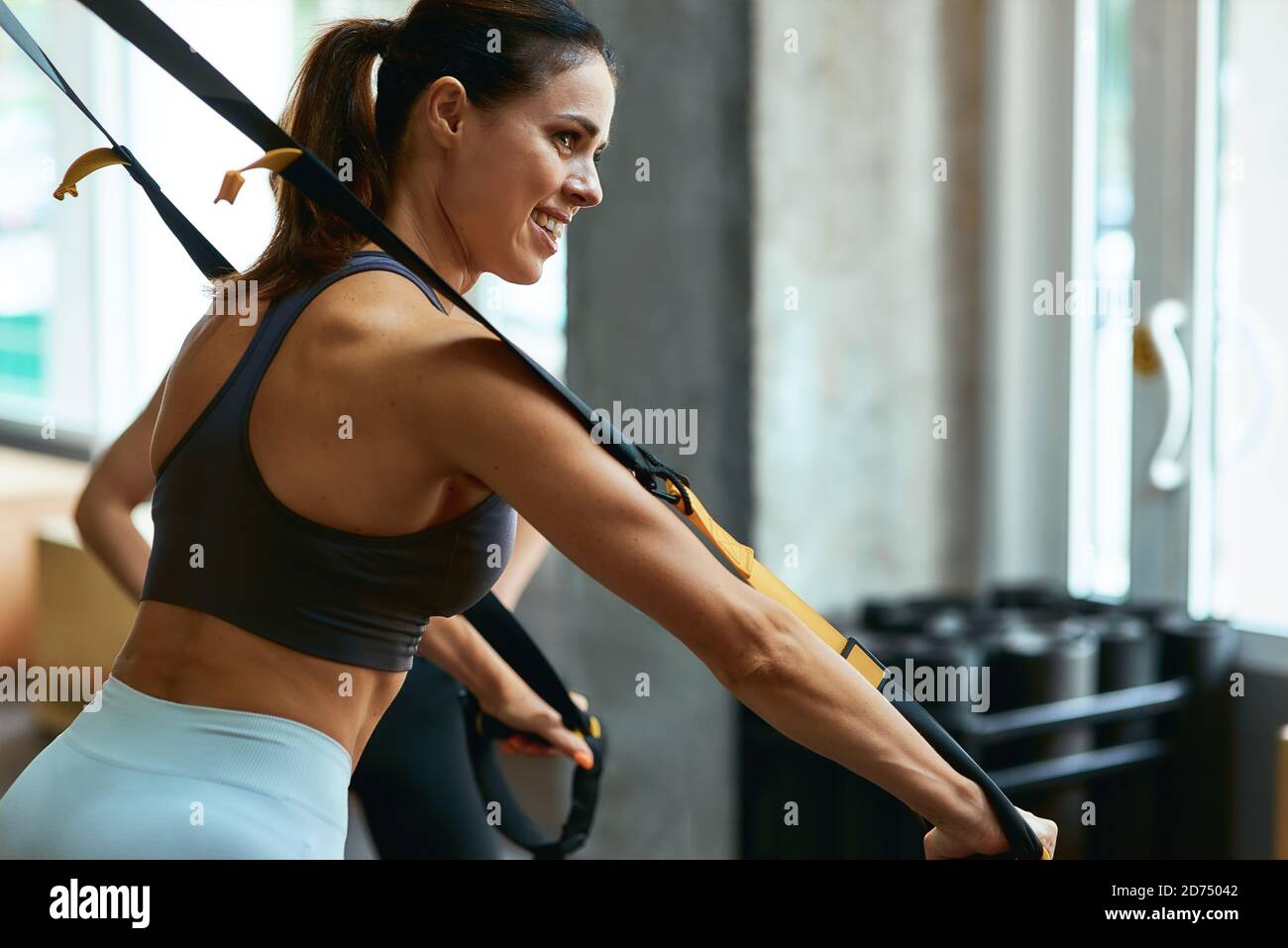 Seitenansicht einer jungen, schönen Frau in Sportbekleidung, die im Fitnessstudio TRX-Workout macht und mit Fitnessbändern trainiert. Sport, Training, Wellness und gesunder Lebensstil Stockfoto
