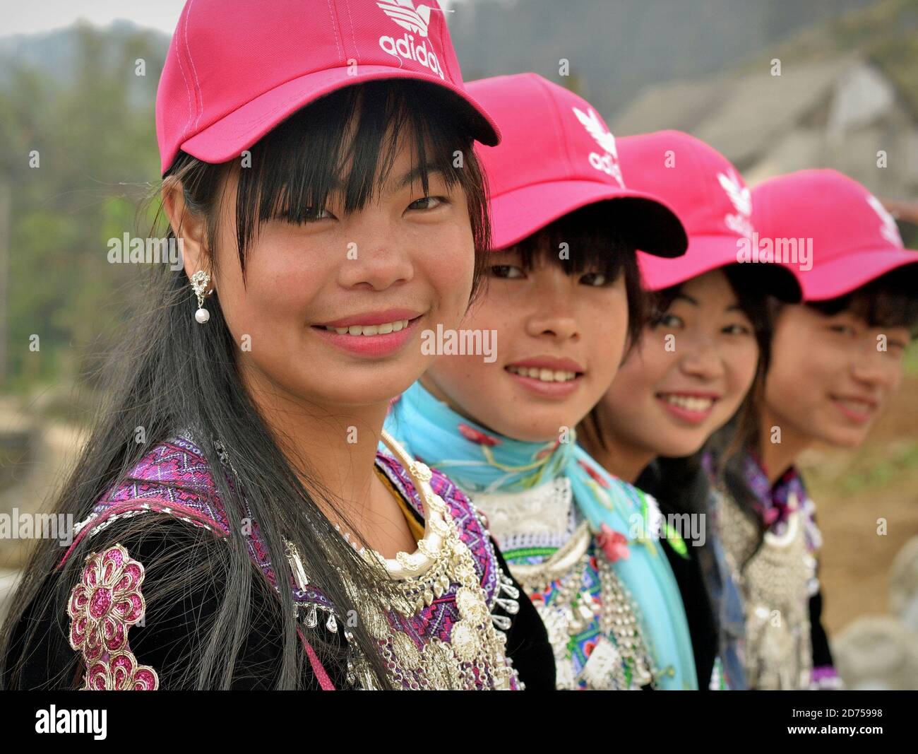 Vier vietnamesische Hmong Hill-Tribe Teenager-Mädchen sitzen in einer Reihe, tragen moderne rosa Baseballmützen und traditionelle Tribal-Kleidung, posieren zusammen für die Kamera. Stockfoto