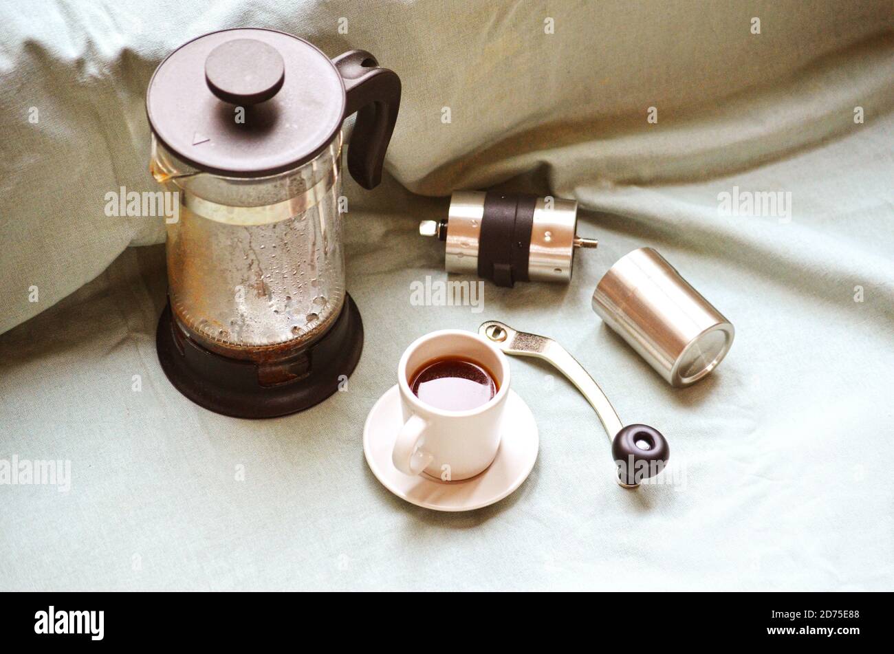 Französische Presse, Kaffeetasse mit dunklem heißem Espresso auf Untertasse und kleine Kaffeemühle aus Metall auf hellgrünem Stofftuch. Stillleben. Herstellung von frischem Coff Stockfoto