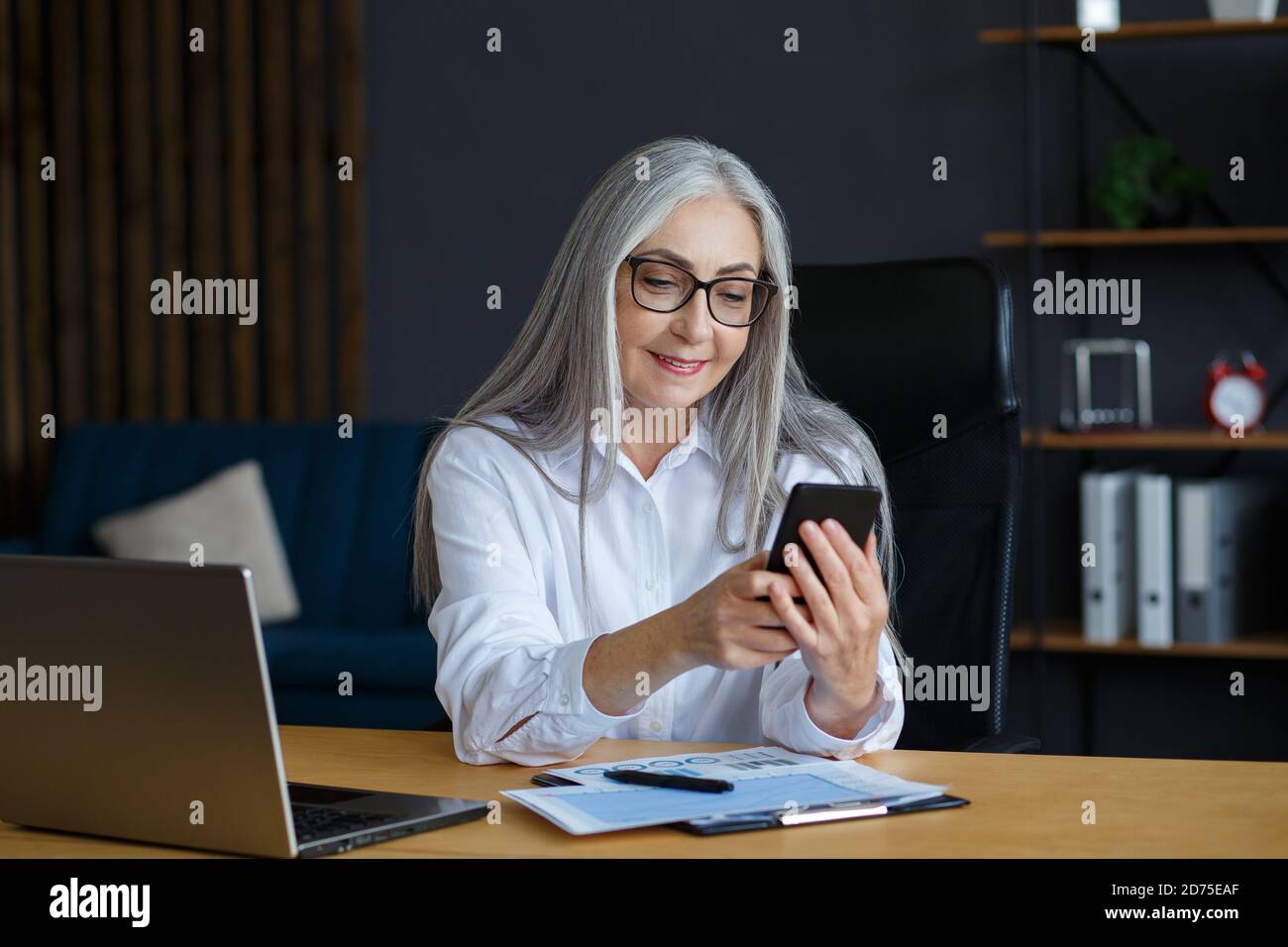 Ältere grauhaarige schöne Frau mit Smartphone und lächeln. Glückliche Geschäftsfrau mit Handy-Apps, SMS-Nachrichten, Surfen im Internet Stockfoto
