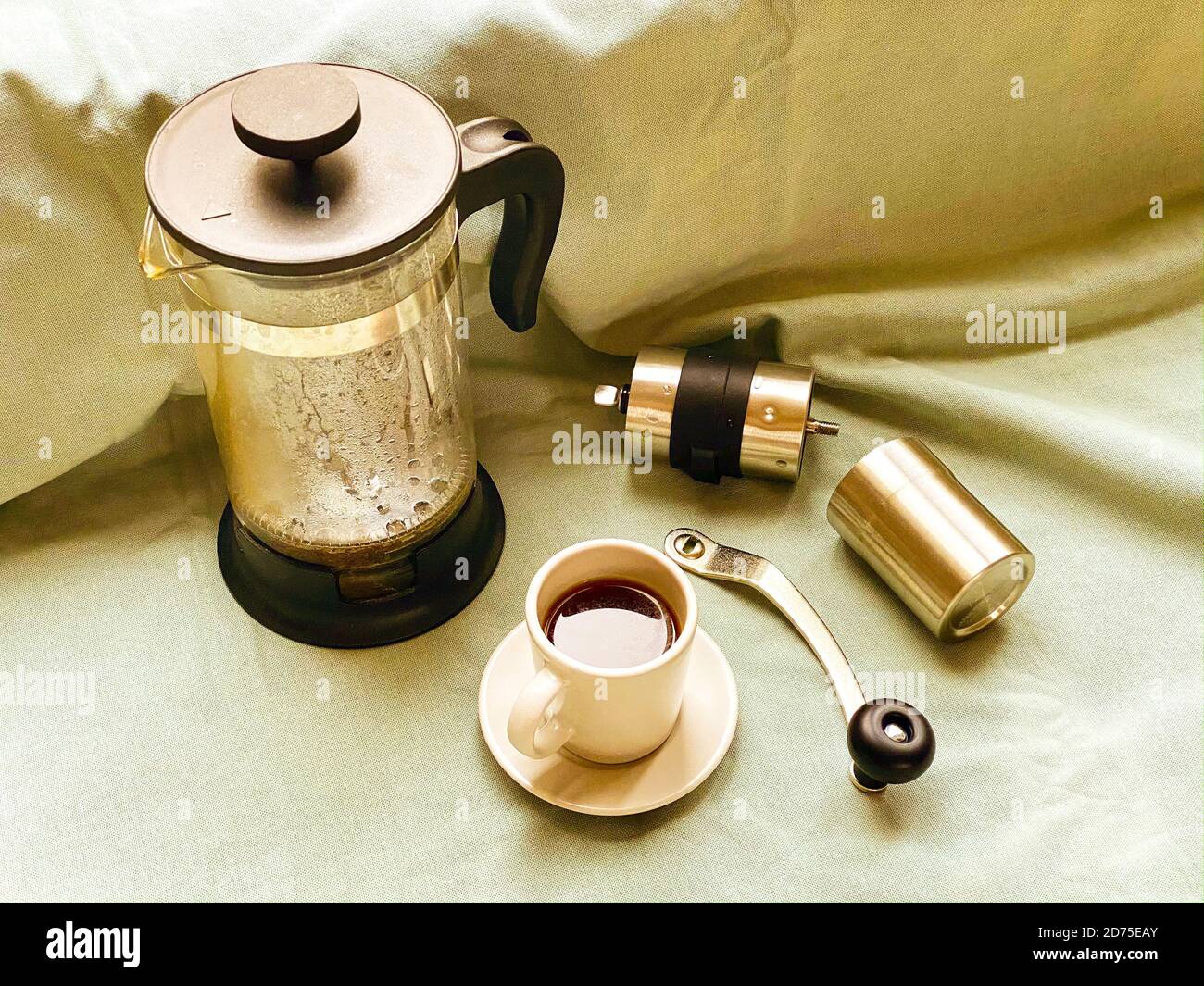 Französische Presse, Kaffeetasse mit dunklem heißem Espresso auf Untertasse und kleine Kaffeemühle aus Metall auf hellgrünem Stofftuch. Stillleben. Herstellung von frischem Coff Stockfoto