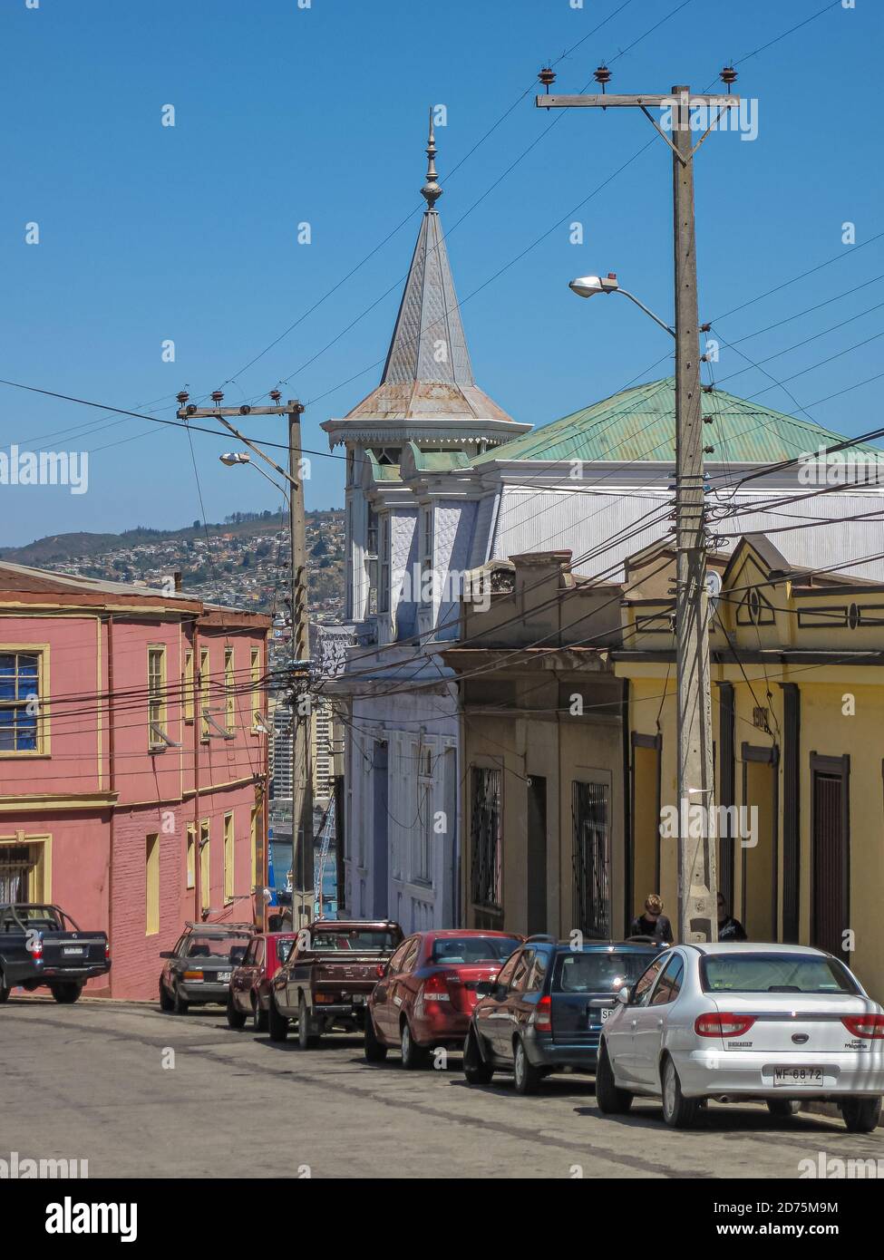 Valparaiso, Chile - 8. Dezember 2008: Straße der unteren Klasse mit ein paar Häusern, eines mit Turm, und Autos vor dem blauen Himmel geparkt. Stockfoto