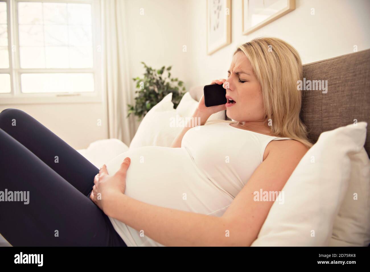 Schwangere Frau auf dem Bett zu Hause mit Kontraktionen halten Mobiltelefon Stockfoto