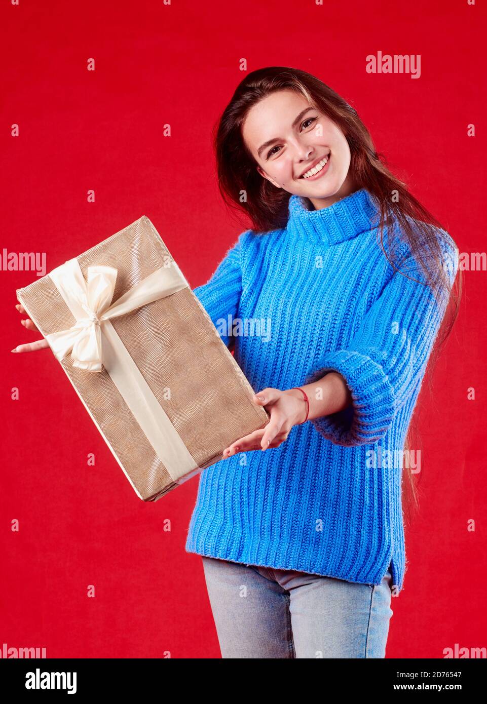 Mädchen in einem blauen Pullover hält ein Geschenk in den Händen auf einem roten Hintergrund. Stockfoto