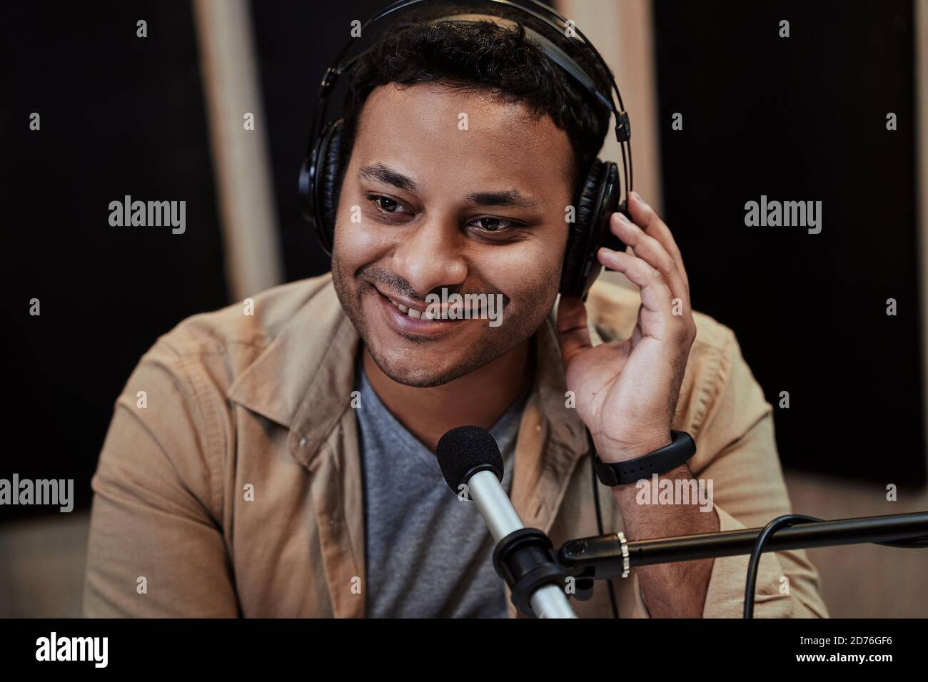 Nahaufnahme von glücklichen jungen männlichen Radiomoderator lächelnd beiseite während der Übertragung im Studio, mit Mikrofon und Kopfhörer Stockfoto