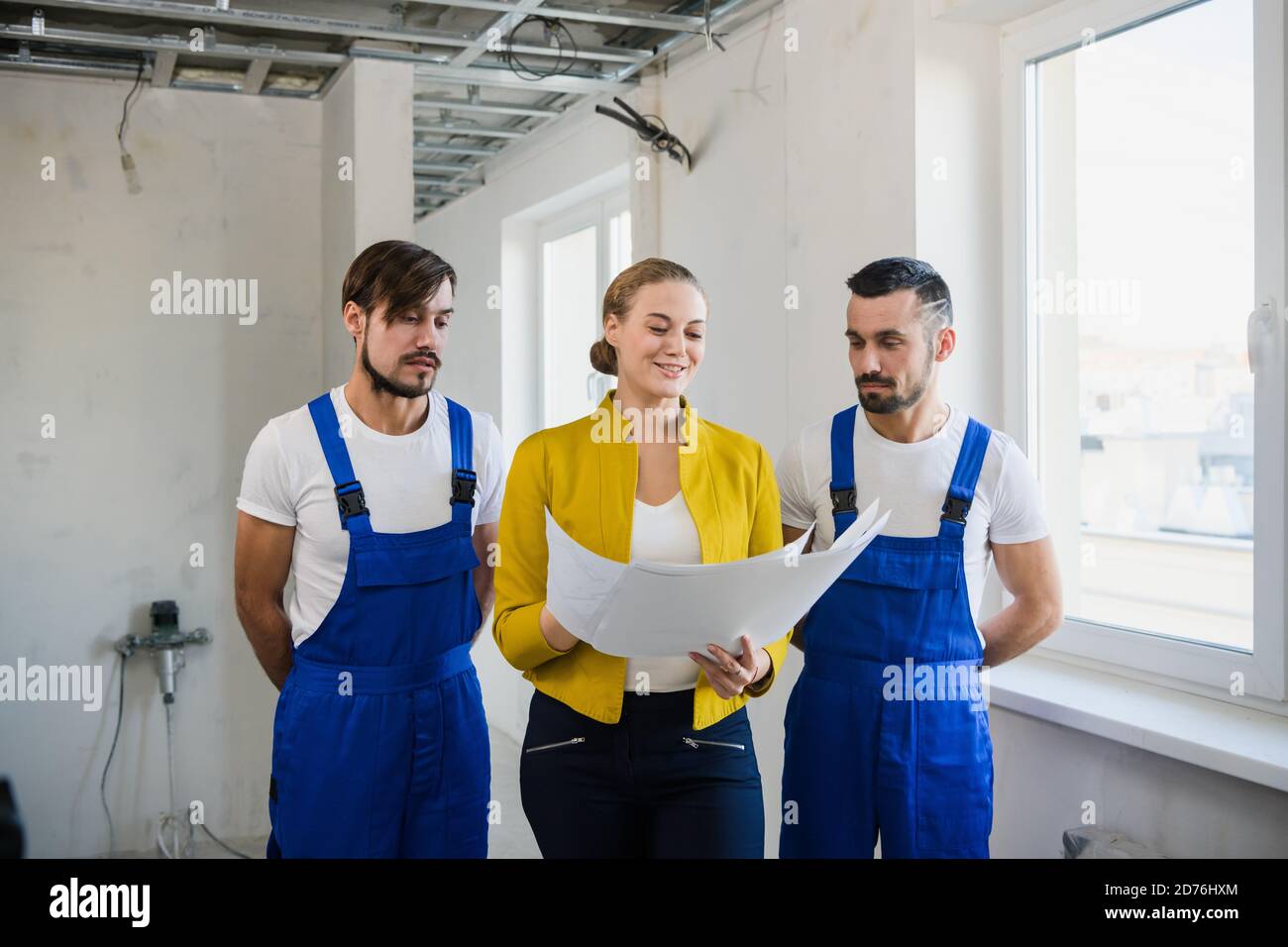 Eine Frau mit einem Hausplan in ihren Händen ist Im Gespräch mit zwei Bauherren in Overalls Stockfoto