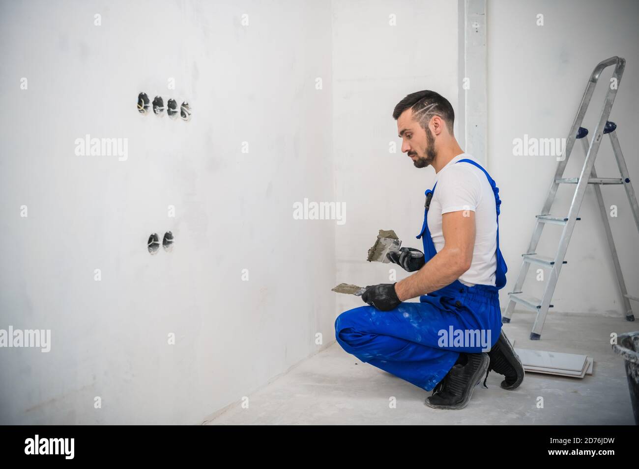 Ein Baumeister in Overalls verwendet eine Kelle, um die zu decken Wand mit Zement Stockfoto