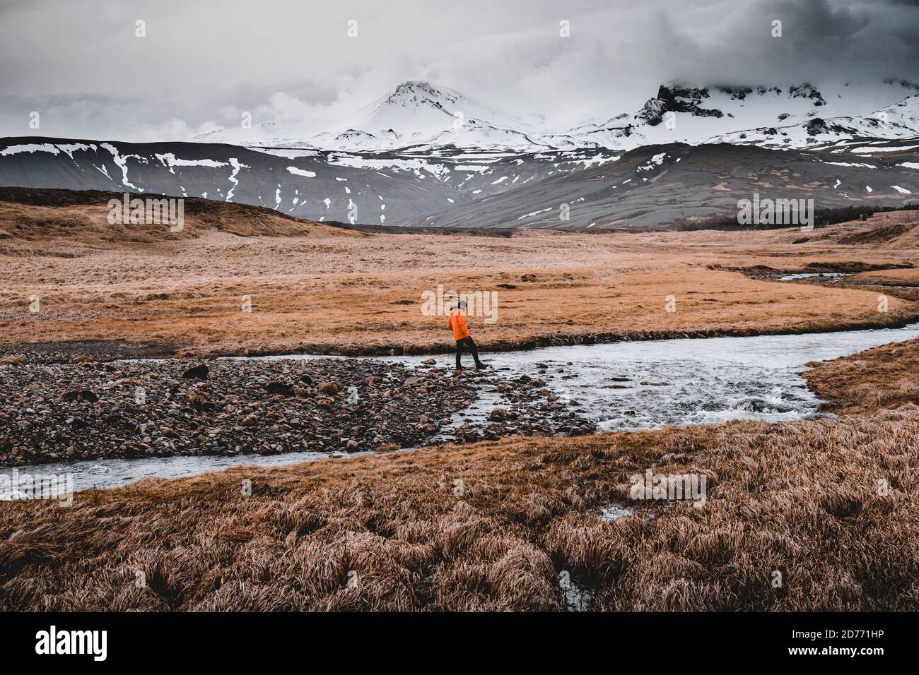 Abenteuerlicher Mann, der auf Felsen über eine flache Flussgegend läuft und hüpft, Eis schmilzt auf einem Fluss in den Bergen, mit Eisberg im Hintergrund Stockfoto