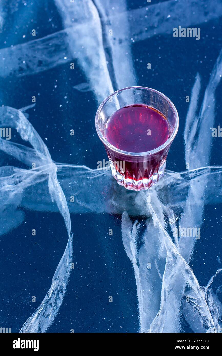 Ein Glas mit roter Tinktur steht auf dem Eis des Sees. Alkohol im Glas und Eis mit schönen tiefen Rissen. Vertikal. Stockfoto