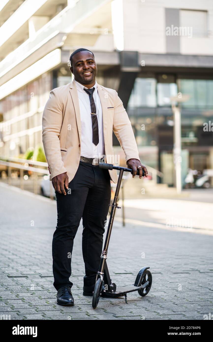 Glücklicher afroamerikanischer Geschäftsmann steht mit Roller vor dem Firmengebäude. Stockfoto