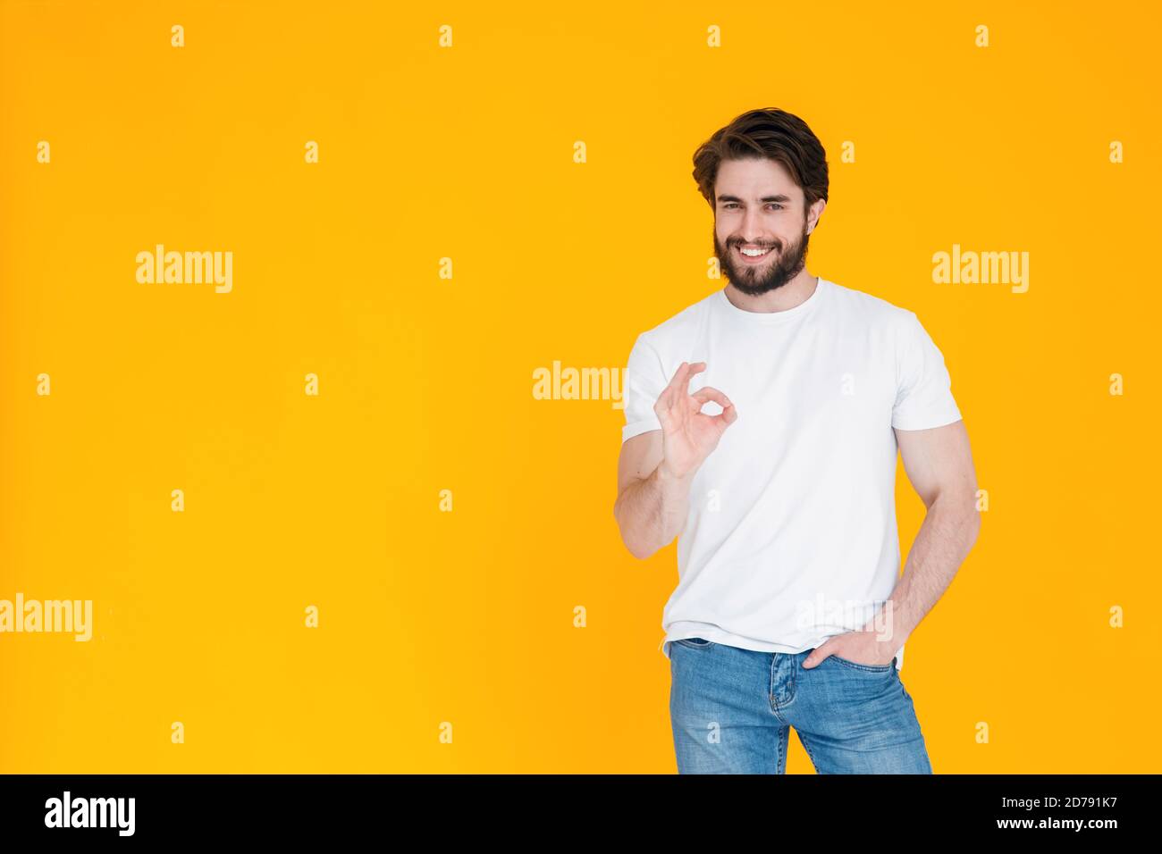Ein glücklicher bärtiger junger Mann in einem weißen T-Shirt Und blaue Denim-Hosen lächeln an der Kamera und zeigen Geste OK Körpersprache alles wird in Ordnung sein auf einem Stockfoto