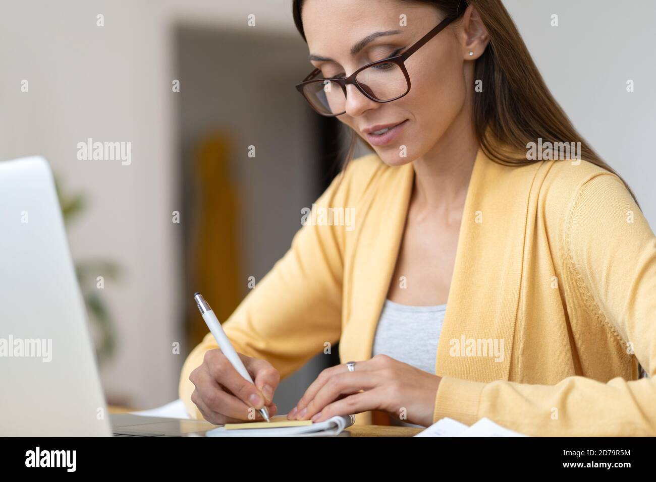 Fokussierte Geschäftsfrau in Brillen tragen gelbe Strickjacke macht eine Notiz in Notebook. Office weibliche Manager aus der Ferne arbeitet von zu Hause während der zweiten w Stockfoto