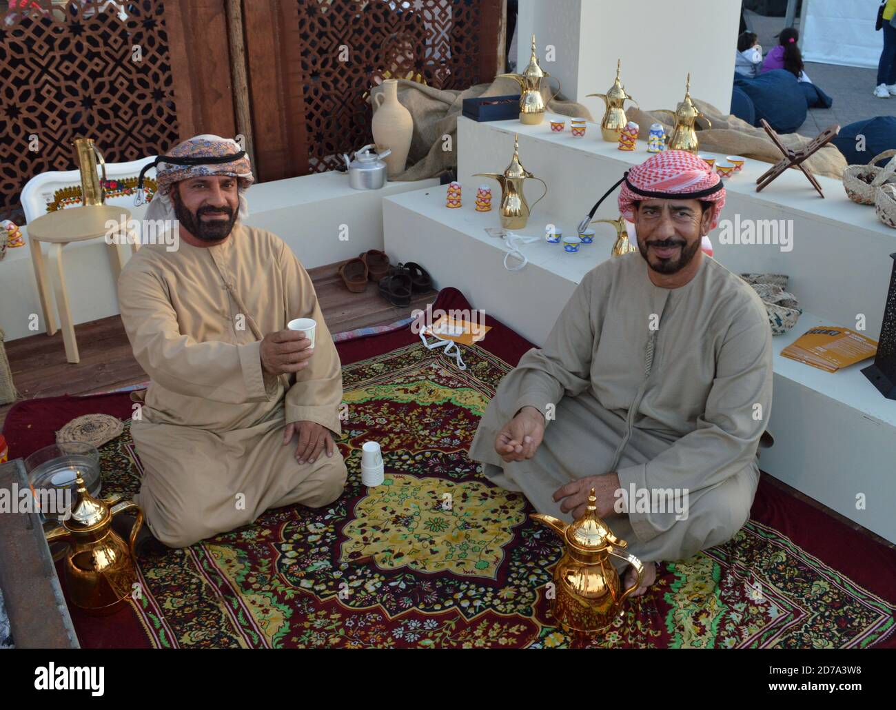 Zwei arabische Männer in traditionellen Kleidern und Kopfbedeckungen trinken Arabisch Kaffee zusammen und lächeln auf der Kamera, wenn sie sitzen Der farbenfrohe orientalische Teppich Stockfoto