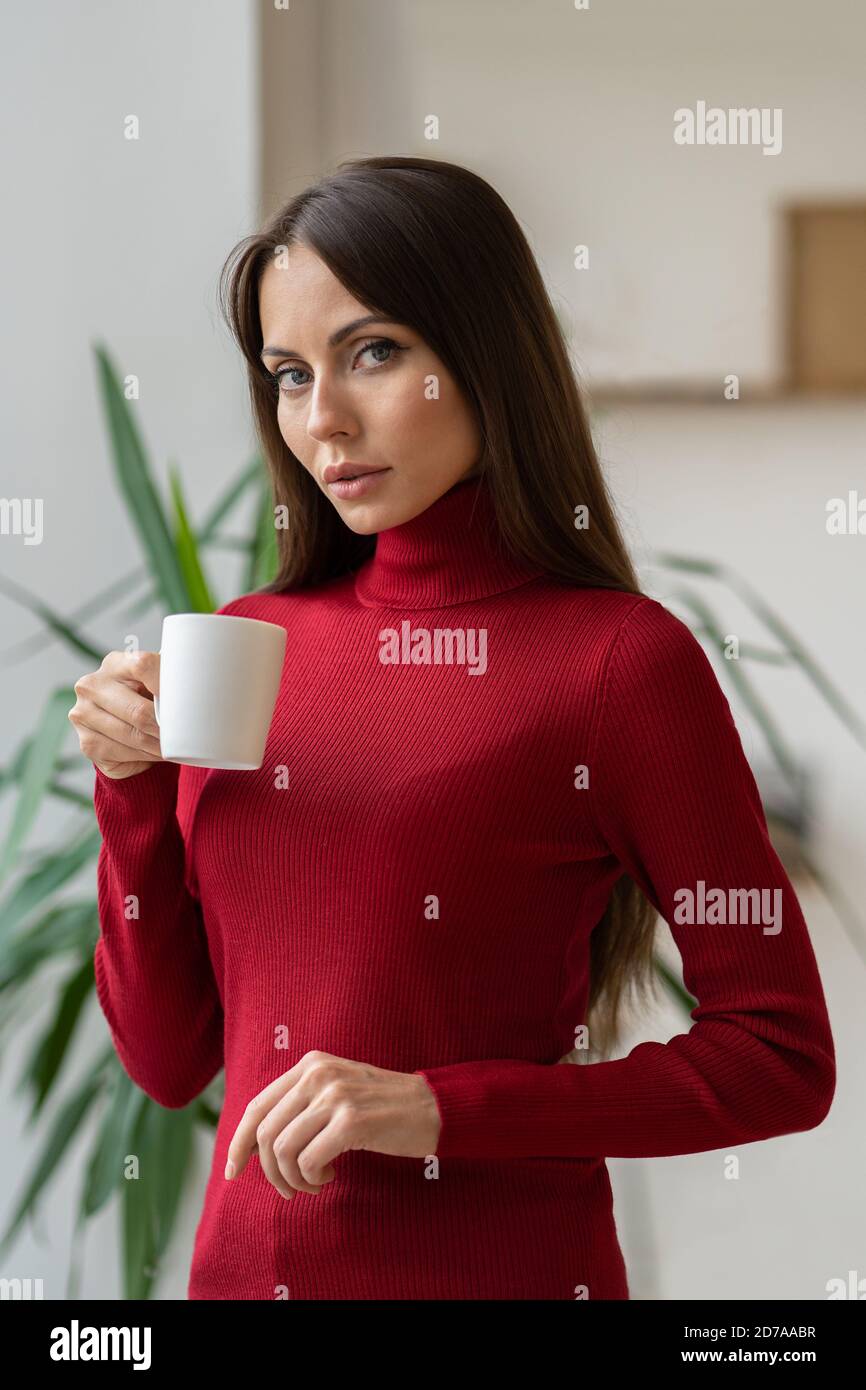 Nachdenkliche kaukasische Frau trägt roten Rollkragen, posiert, Blick durch die Kamera, hält weiße Tasse, trinken Kaffee oder Tee. Stockfoto