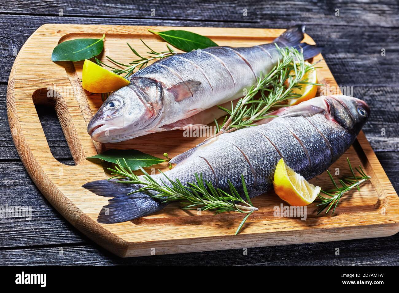 Seabass frischer roher Fisch auf einem Schneidebrett mit frischem Rosmarin, Zitronenkeilen und Lorbeerblatt auf einem Schneidebrett auf einer dunklen Holzba entkaltet und entkalkt Stockfoto