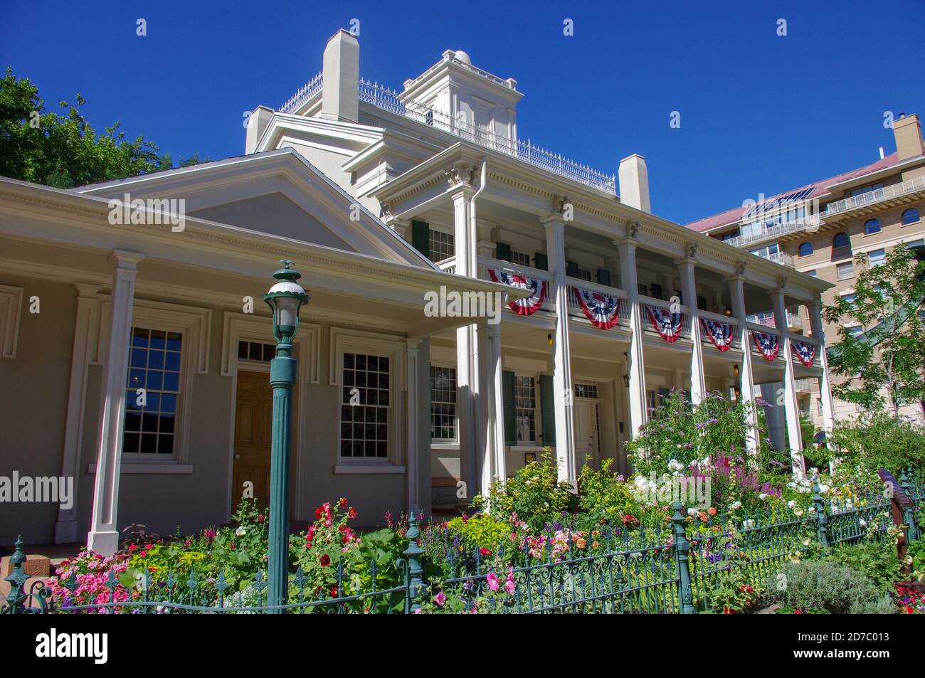 Salt Lake City, UT - 12. Juli 2012; Blumengärten liegt hinter grünem Eisenzaun vor dem Pionier-Holzhaus Bee Hive mit patriotischem Haunting Hangin Stockfoto