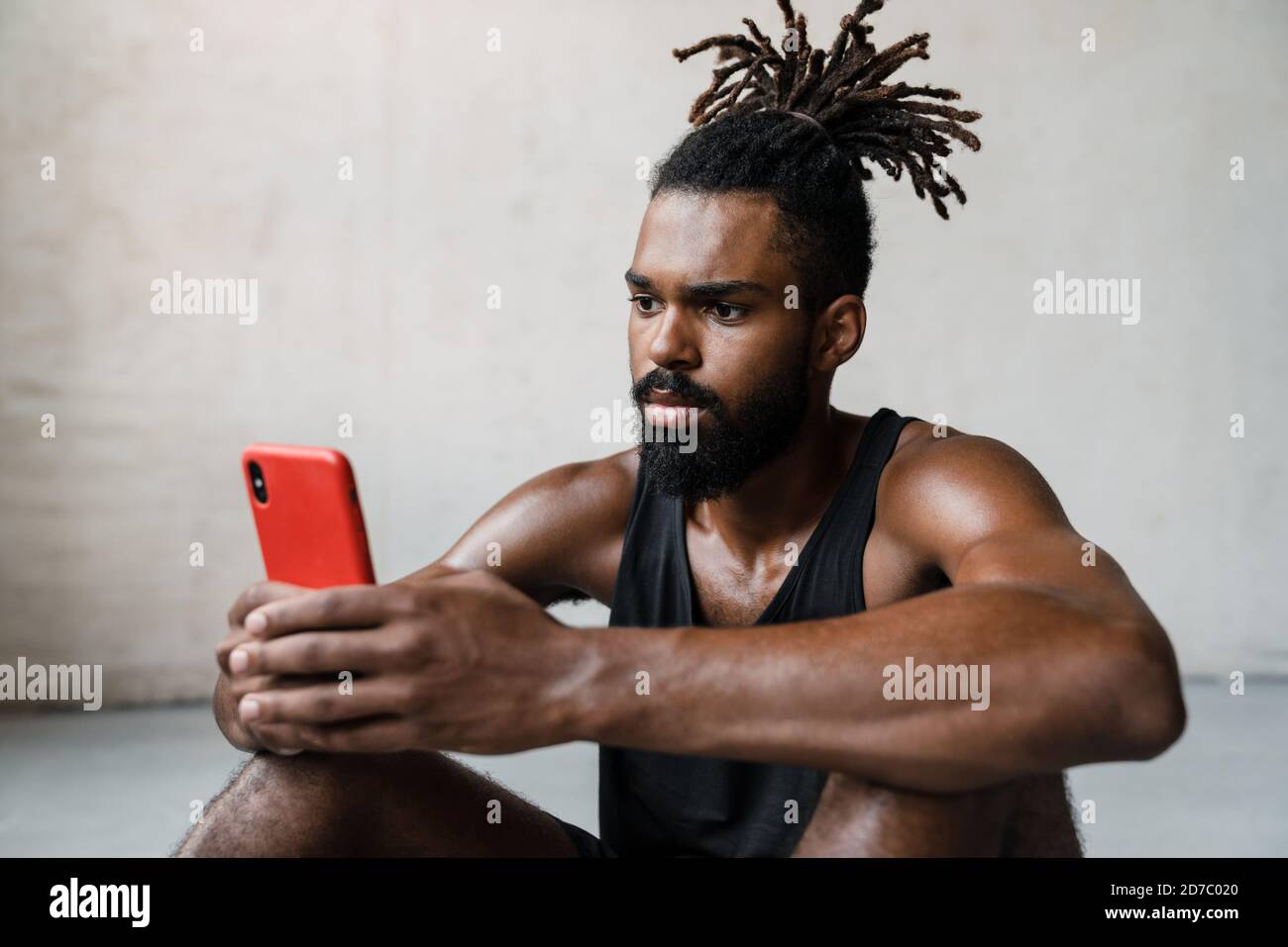 Bild von jungen afroamerikanischen Sportler mit Handy während Drinnen auf dem Boden sitzen Stockfoto