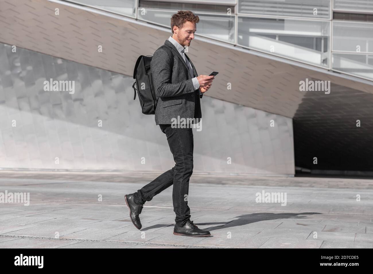 Glücklicher junger Geschäftsmann, der mit dem Telefon unterwegs ist, pendelt zur Arbeit mit einer Rucksack-Tasche auf der Stadtstraße. Geschäftsmann SMS Blick auf Smartphone Stockfoto