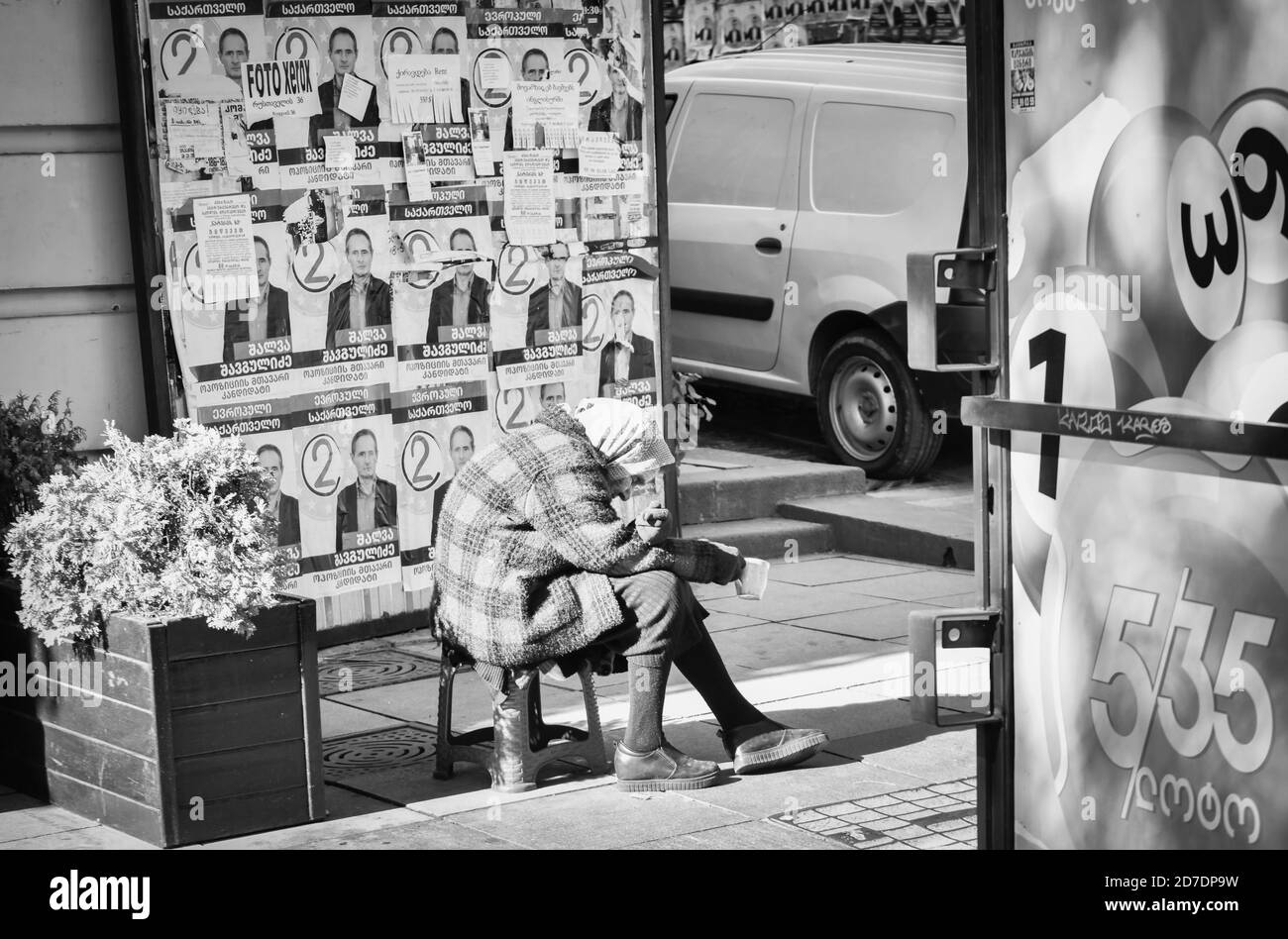 Die alte Frau sitzt auf dem Plastikstuhl und bittet darum, die grüne Tasse mit dem Kopf auf den Boden zu halten. Betteln in großen Städten Konzept. 8. oktober 2020.Tiflis. Georgien Stockfoto