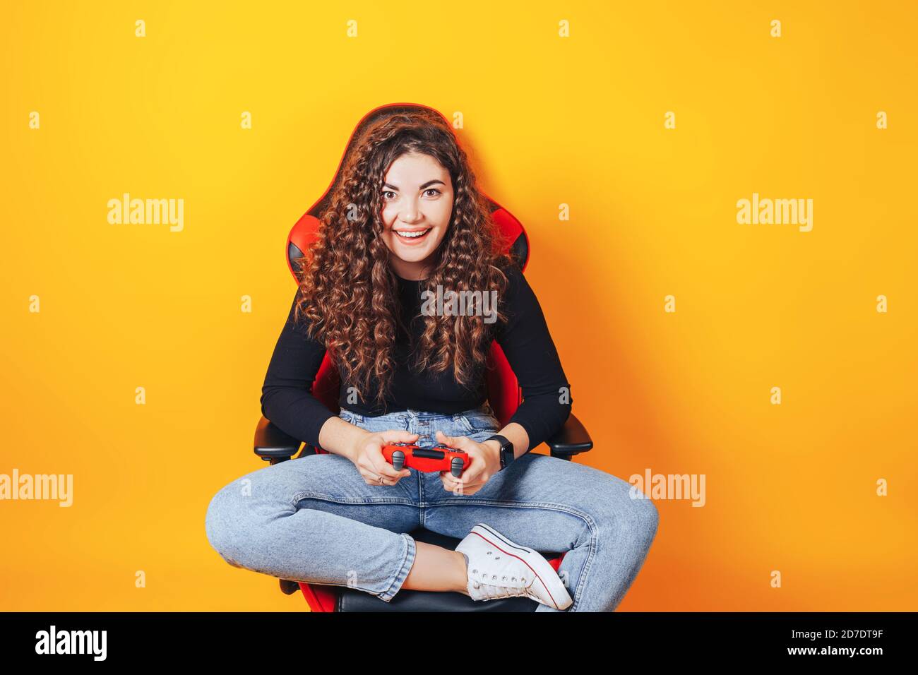 Frau, die hinter einem Spielstuhl in ihren Händen sitzt und rot hält Gamepad auf gelbem Hintergrund Stockfoto