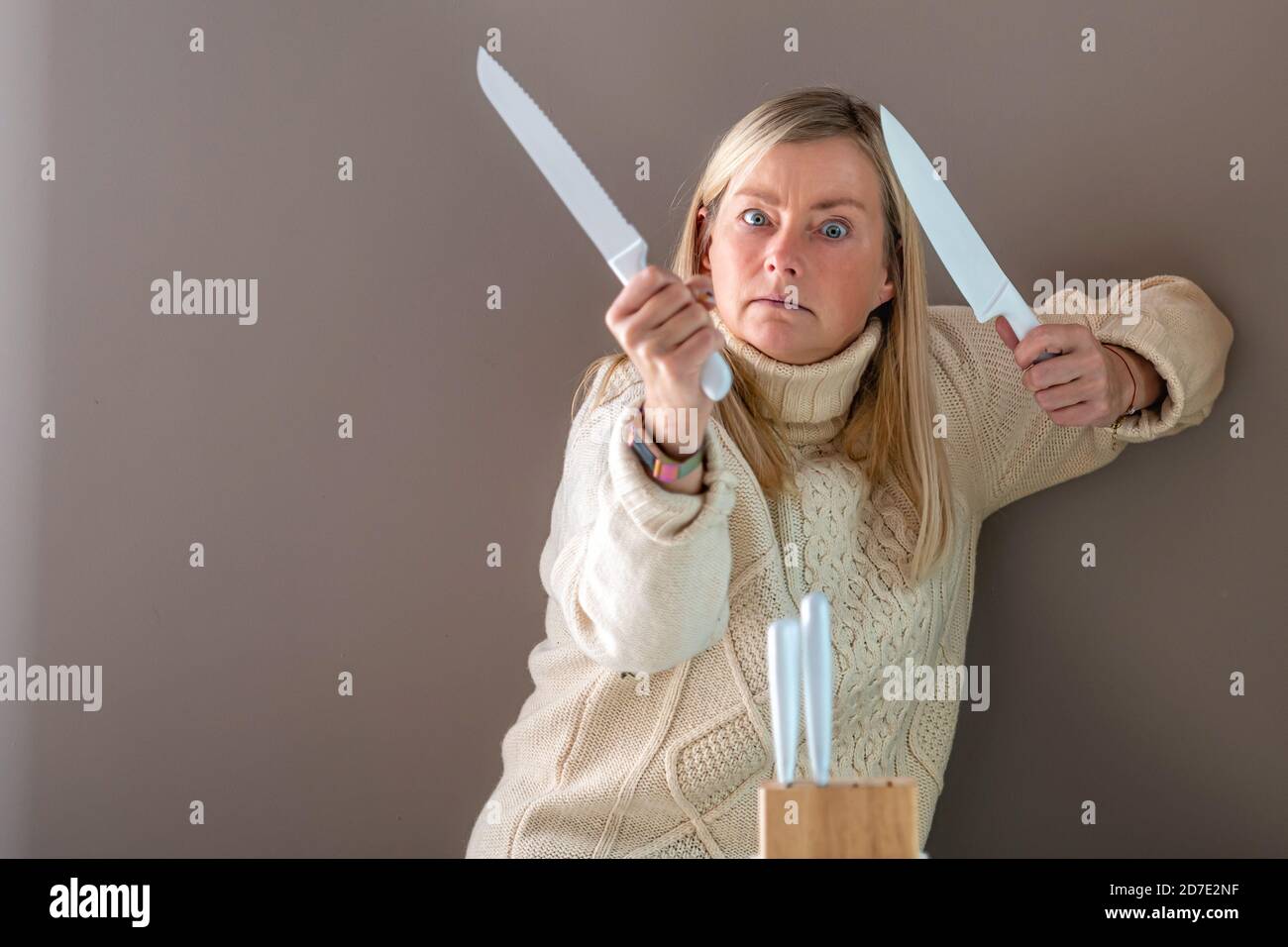 Blonde Frau mit zwei weißen Messern in der Hand, im Vordergrund ein Satz Küchenmesser auf dem Tisch Stockfoto