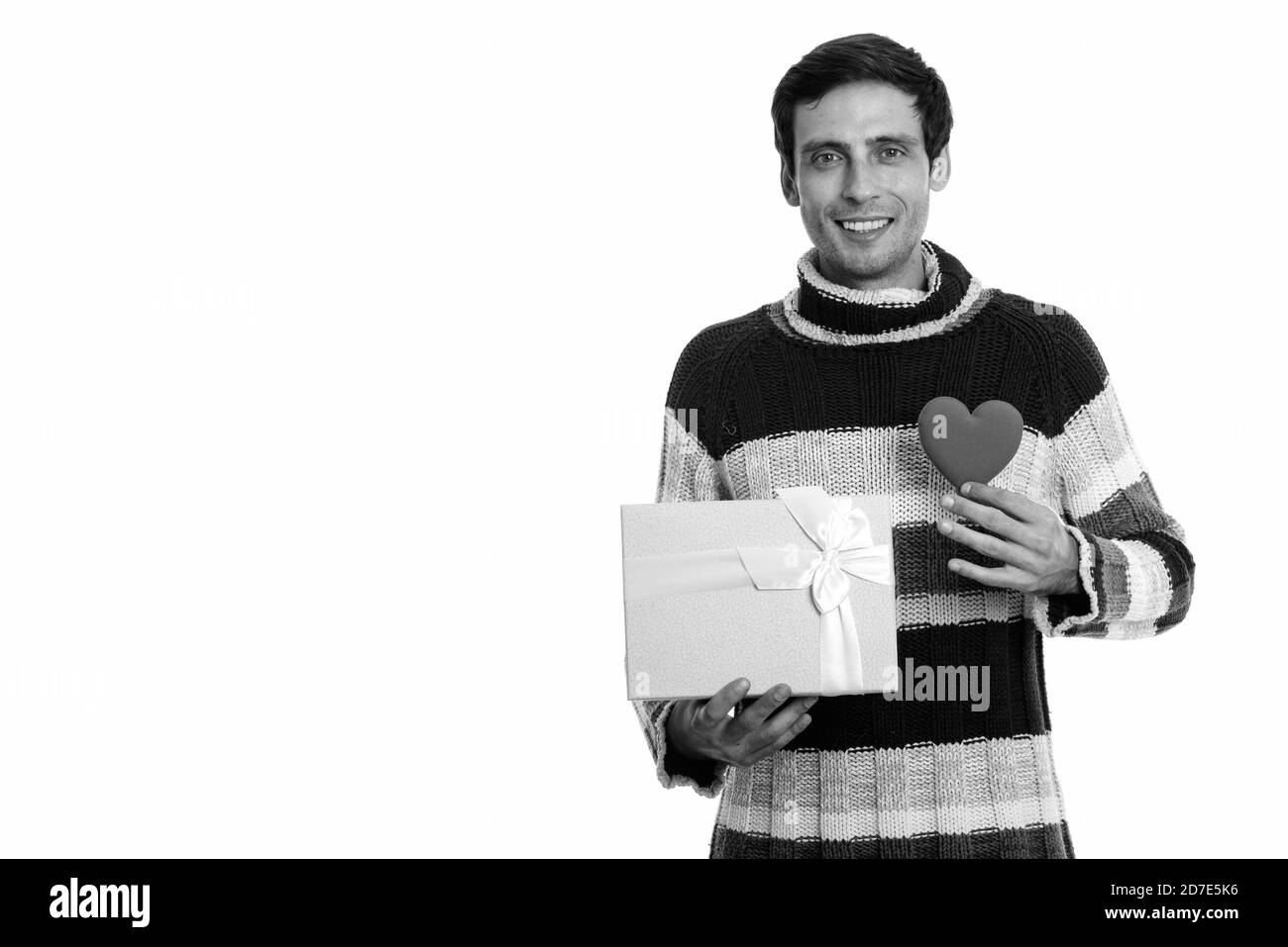 Studioaufnahme eines glücklichen jungen, gutaussehenden Mannes, der beim Halten lächelt Geschenkbox und Herz bereit für Valentinstag Stockfoto